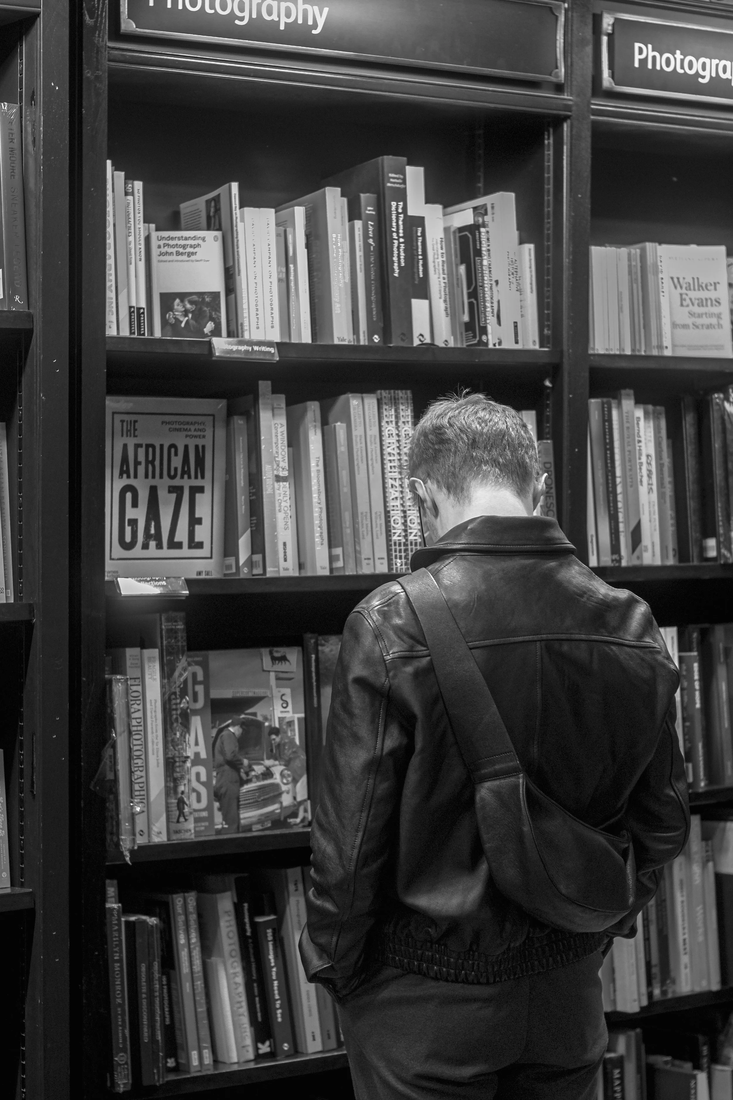 A person with short hair and a leather jacket stands in front of a bookshelf filled with photography books, with the back of their head and shoulders visible.