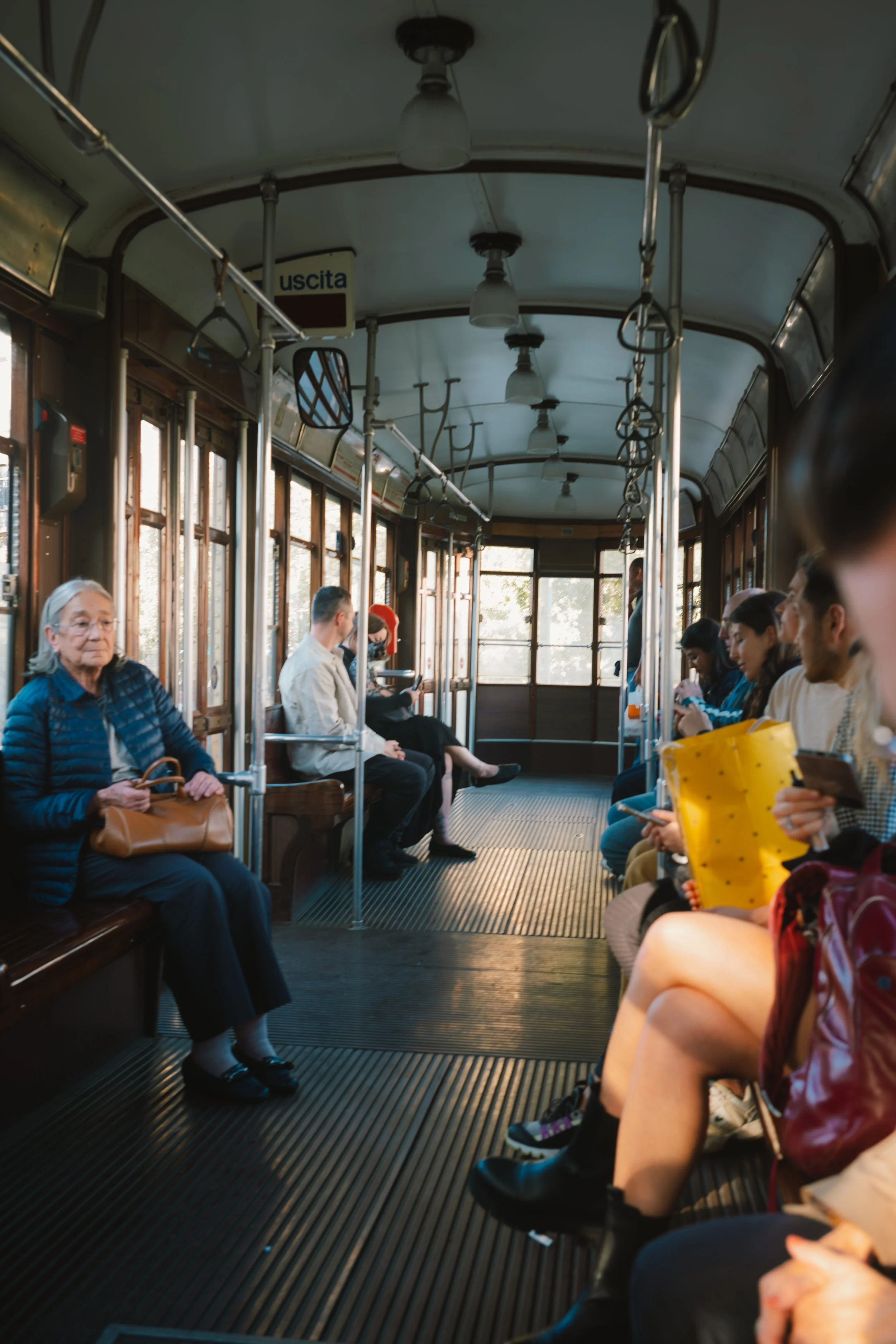 Interior of a vintage tram or train car with passengers seated on wooden benches. Some passengers are wearing masks, and others are looking at their phones. The tram has a curved ceiling with hanging lights and hand straps for standing passengers. An