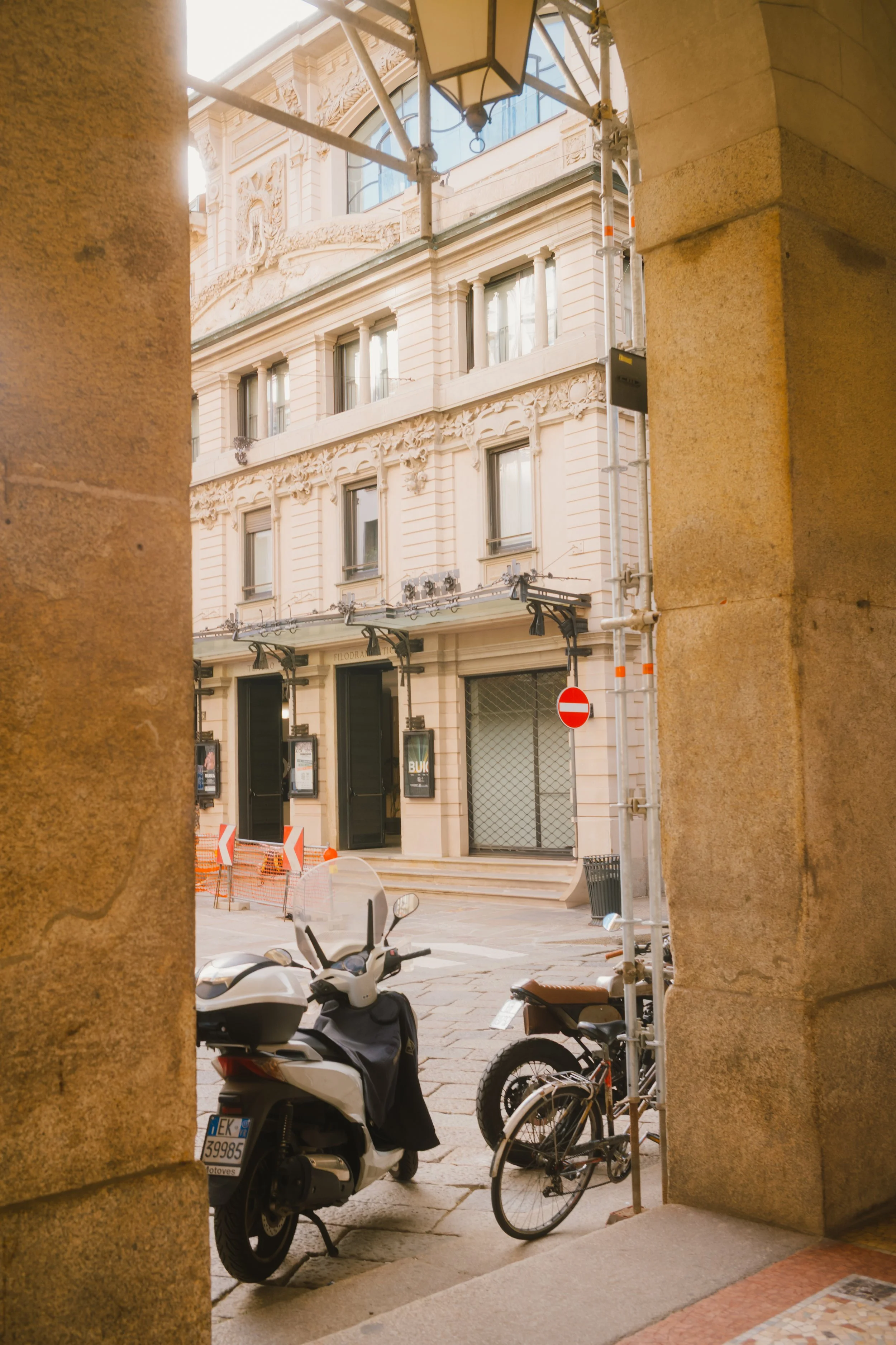 View of a European street scene through a stone archway, featuring parked motorcycles and bicycles, a building with ornate architectural details, and street signs.