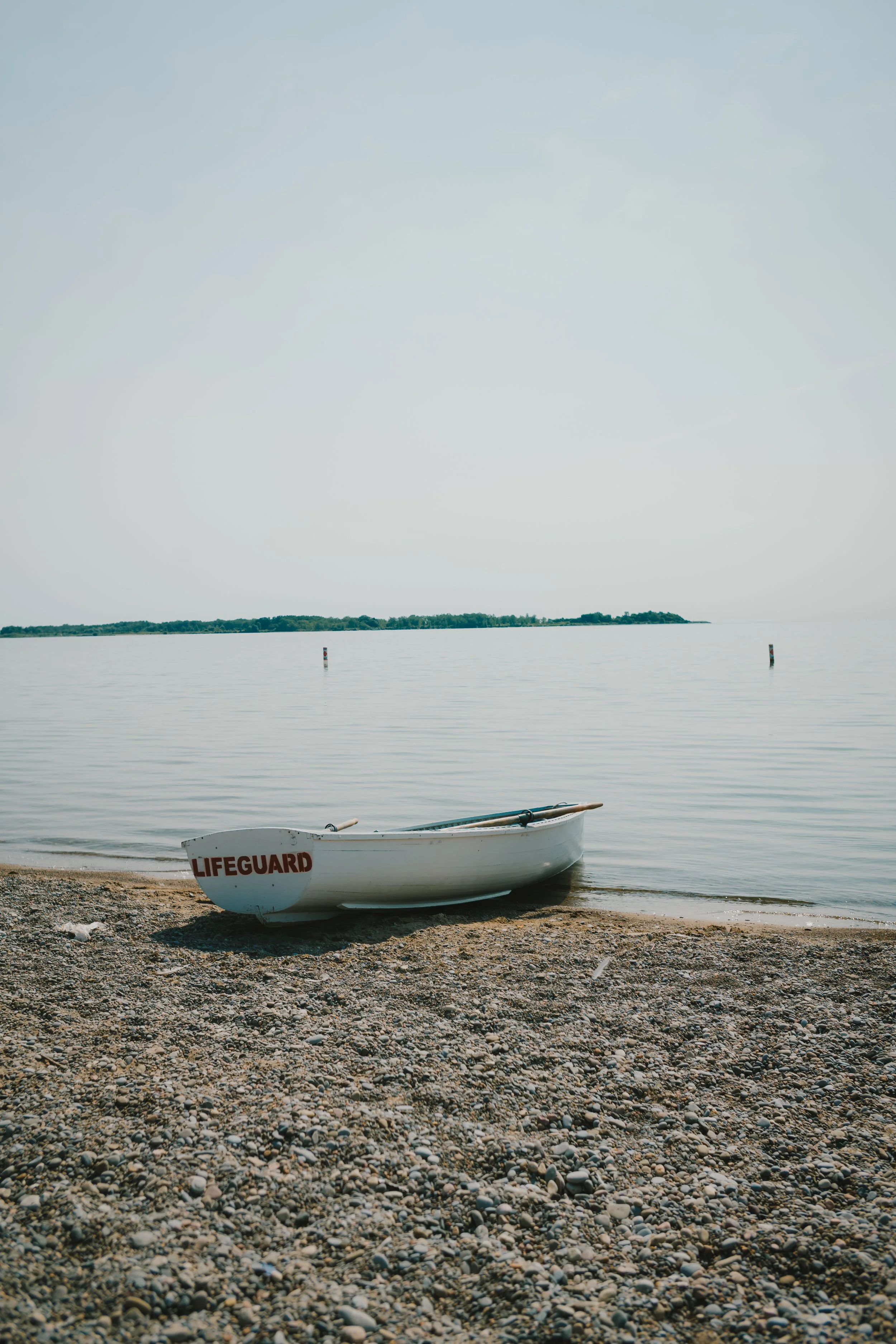 A white lifeguard boat resting on a pebble beach by calm water, with a distant landmass and a cloudy sky in the background.