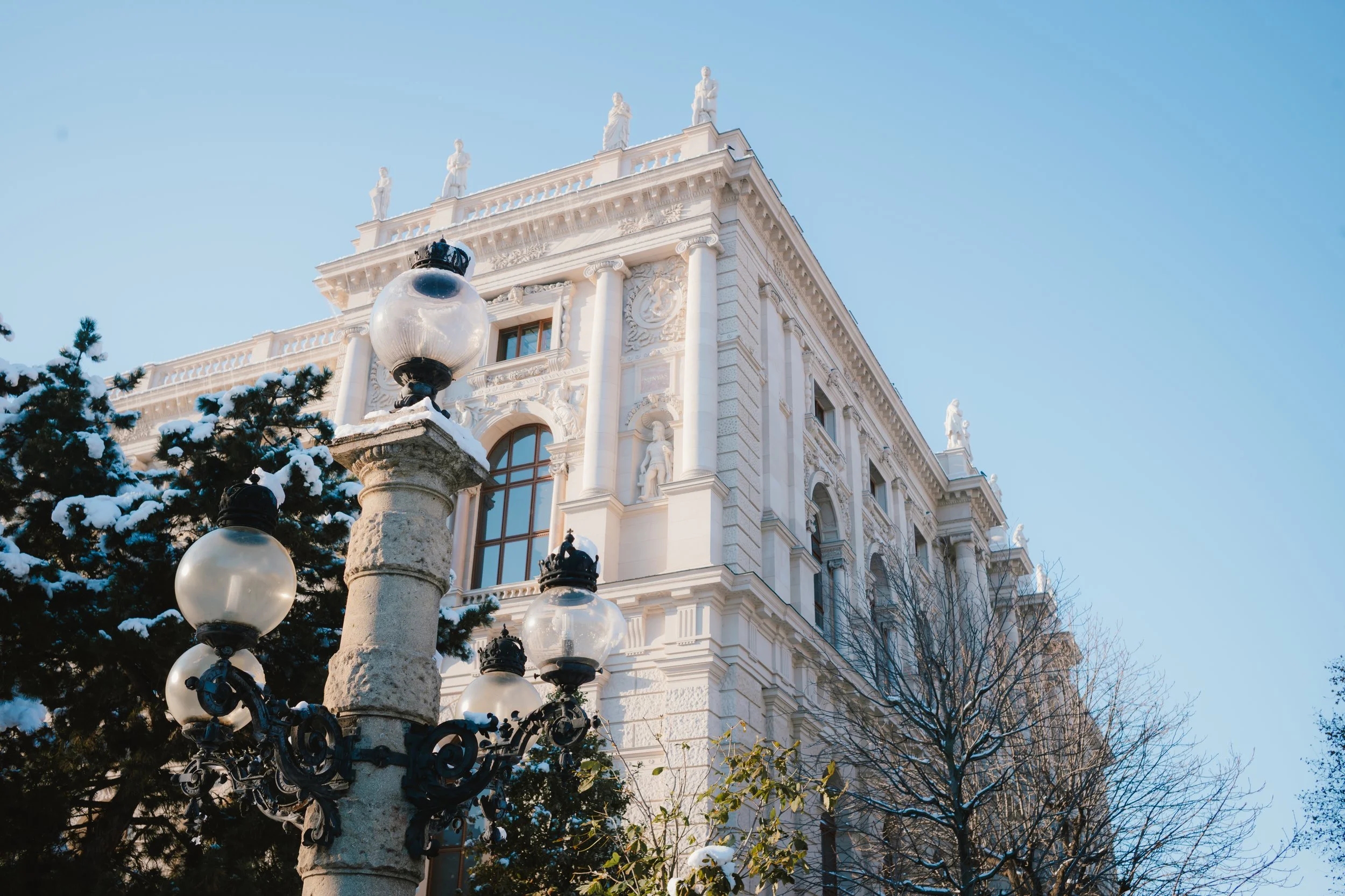 A historic white building with ornate statues on the roof, snow-covered trees, and vintage street lamps in the foreground against a clear blue sky.