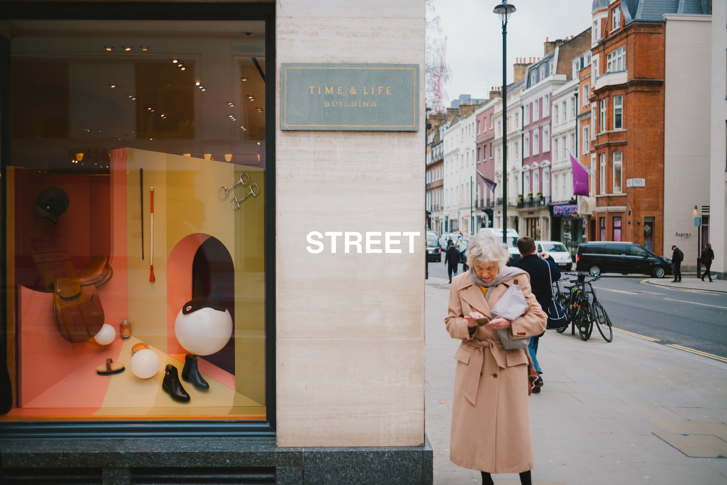 A street scene with a storefront window display on the left and a woman in a tan coat looking at her phone on the right. The window display includes fashion accessories and decorative objects. The background shows colorful buildings, cars, bicycles, 