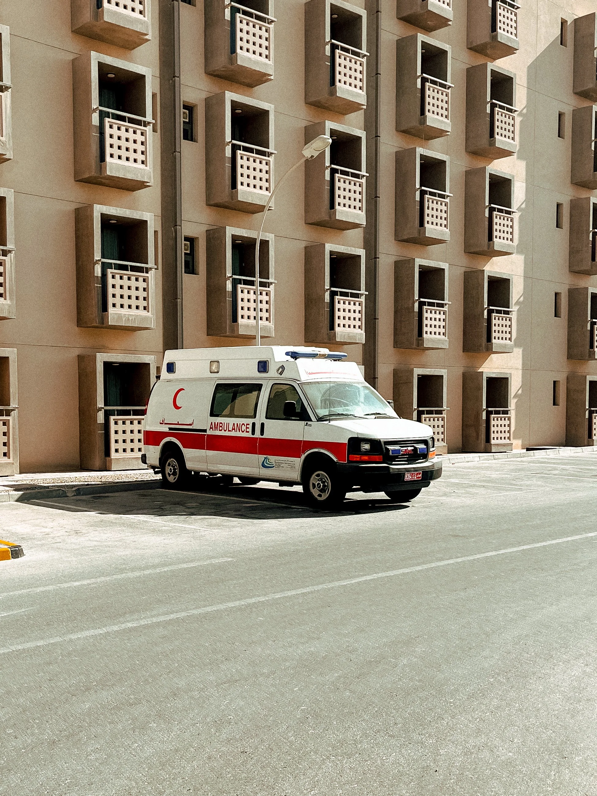 A white ambulance with red stripes and Arabic writing parked on a street in front of a modern apartment building with multiple balconies.