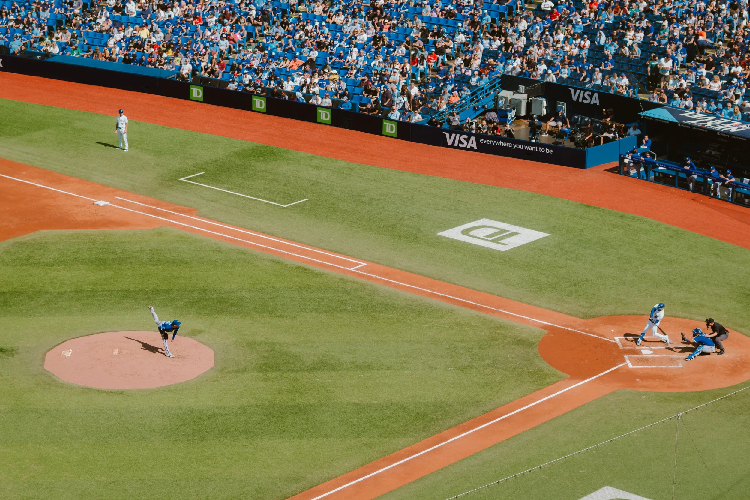 Baseball game in progress with players and audience, focusing on pitcher, batter, and catcher at the home plate, with advertisements on the outfield wall.