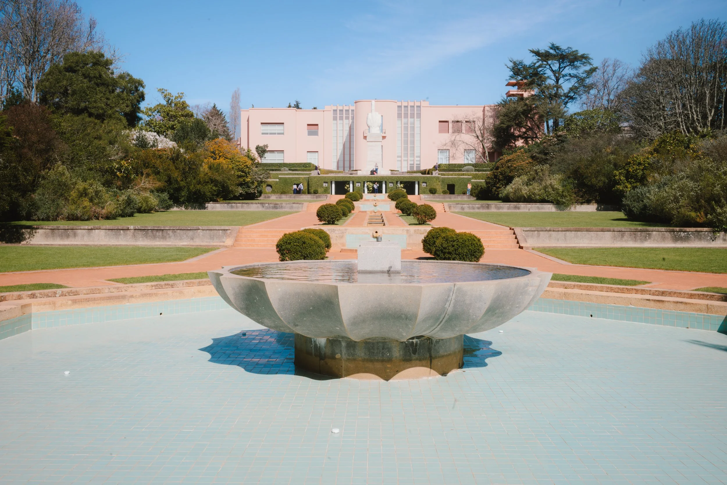 A pink building with a modern architectural design, surrounded by a landscaped garden with trees and shrubs, and a large fountain in the foreground.