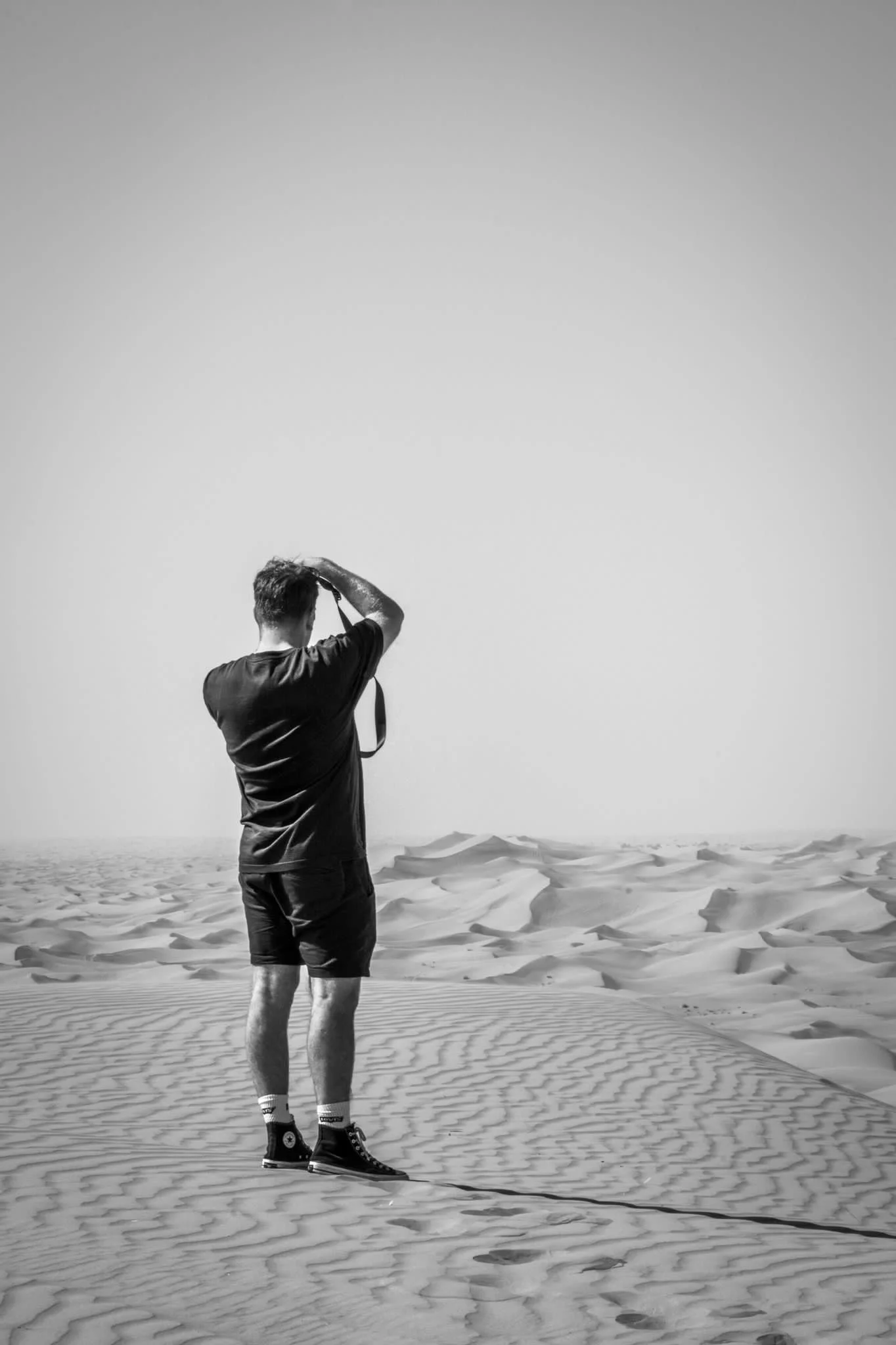 A man standing on a sandy desert with dunes in the background, wearing shorts, a t-shirt, and ice hockey skates, holding a helmet, with footprints visible in the sand.