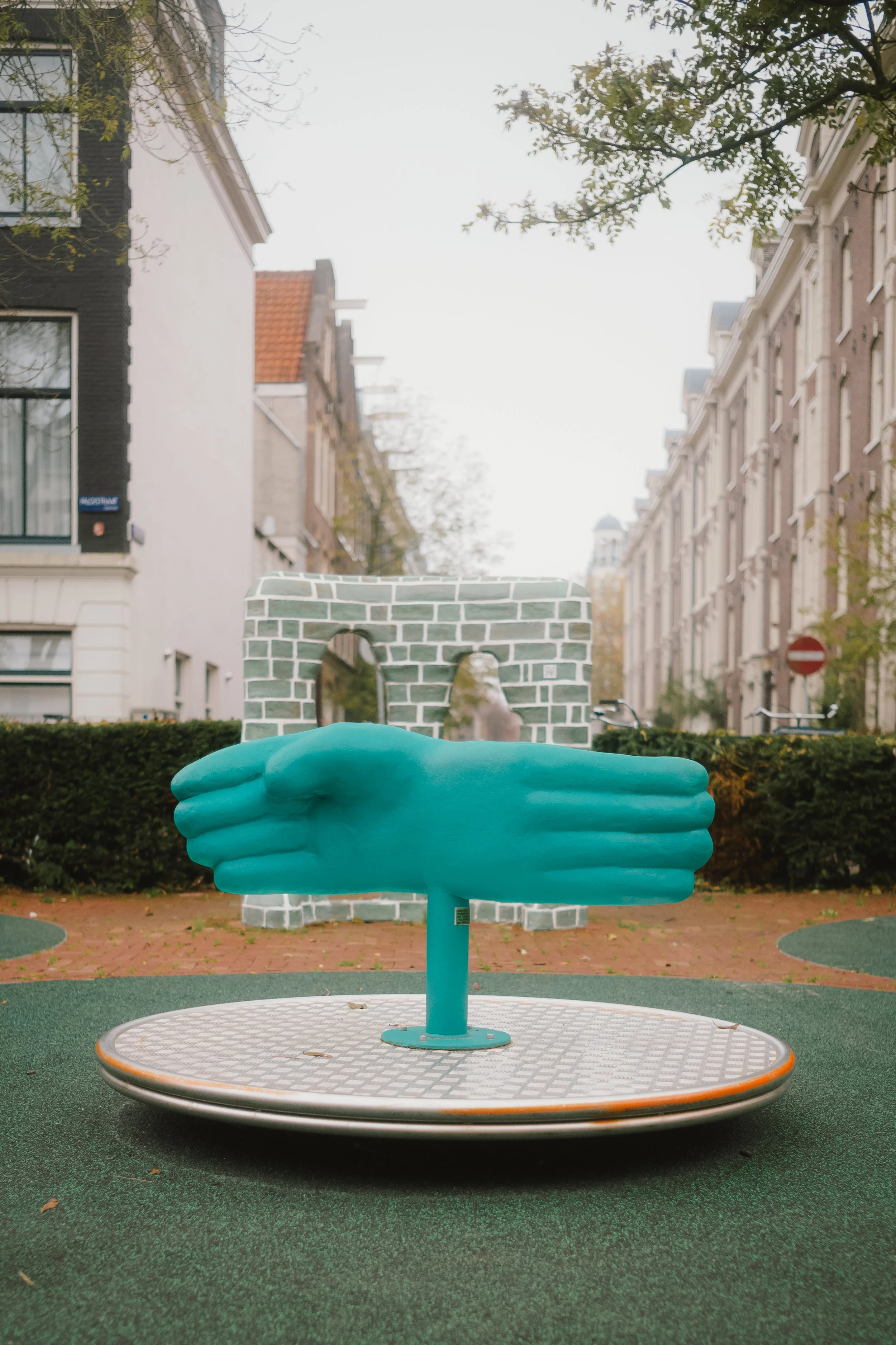 A blue sculpture of two hands holding a brick wall arch, standing on a circular platform in a park with trees and apartment buildings in the background.