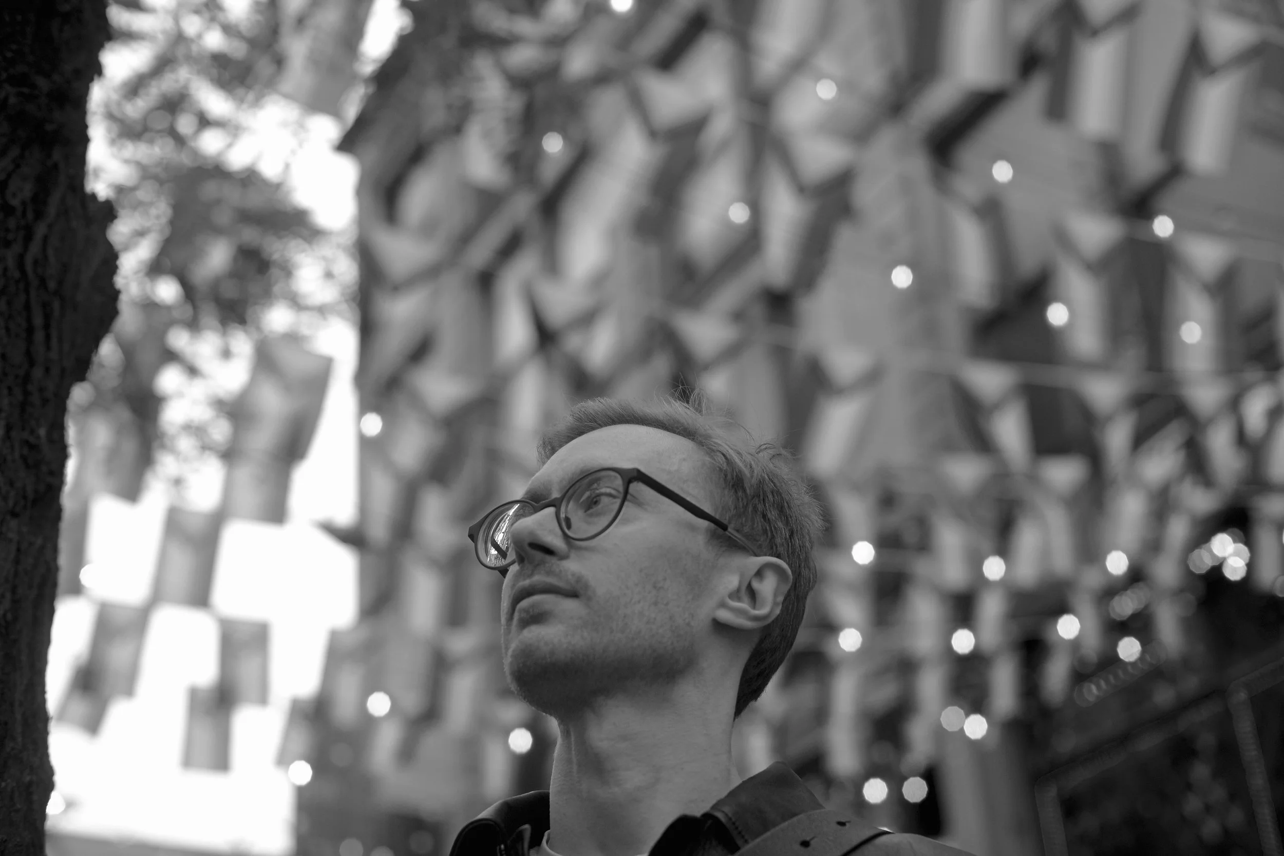 A black-and-white photo of a young man with glasses looking upward in an outdoor urban setting, with large buildings and string lights in the background.