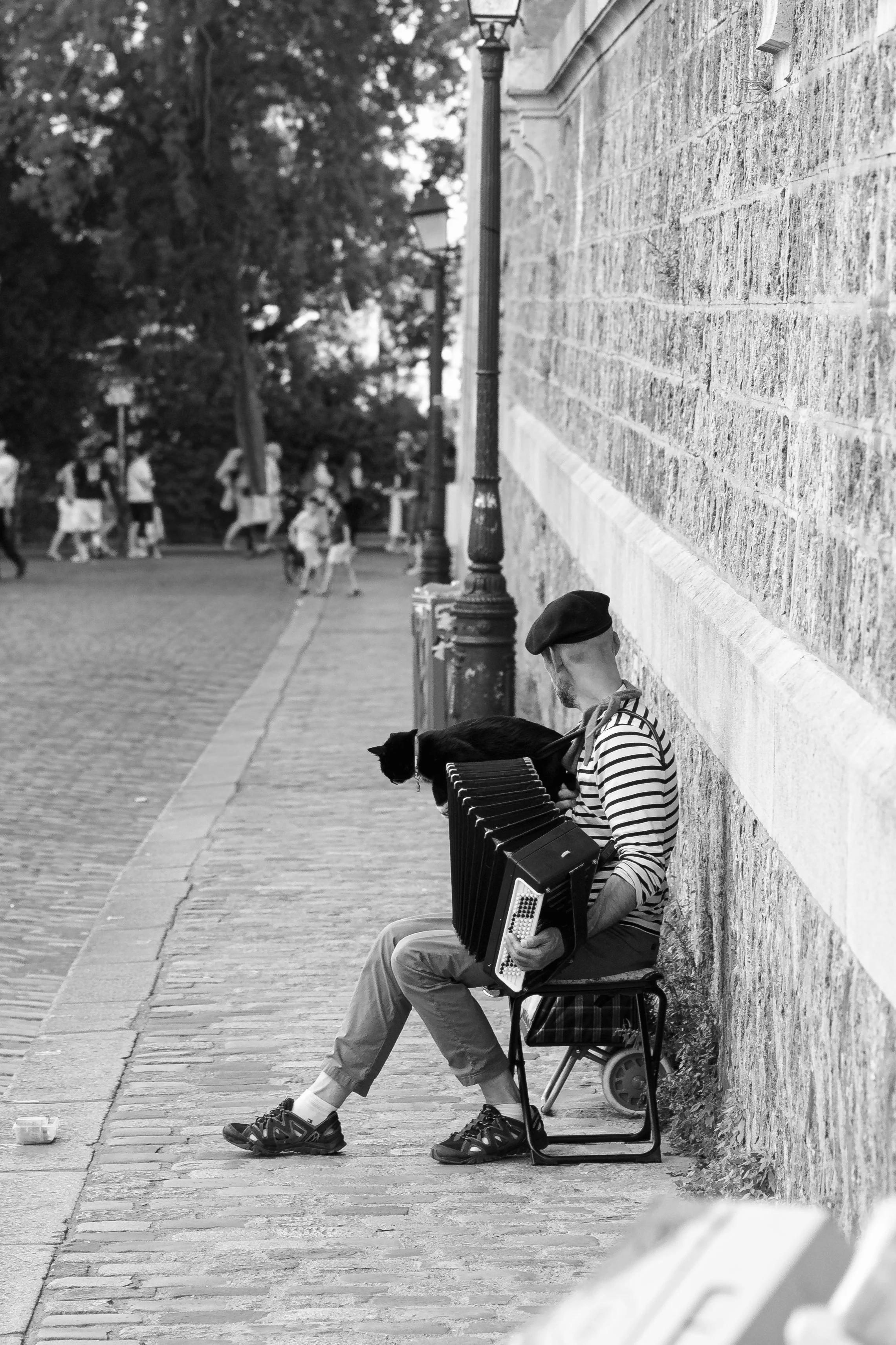 A man sitting on a bench playing an accordion on a city sidewalk, wearing a striped shirt and a beret, with a dog on his lap. In the background, several children are walking along the sidewalk, and old-fashioned street lamps are visible along the str