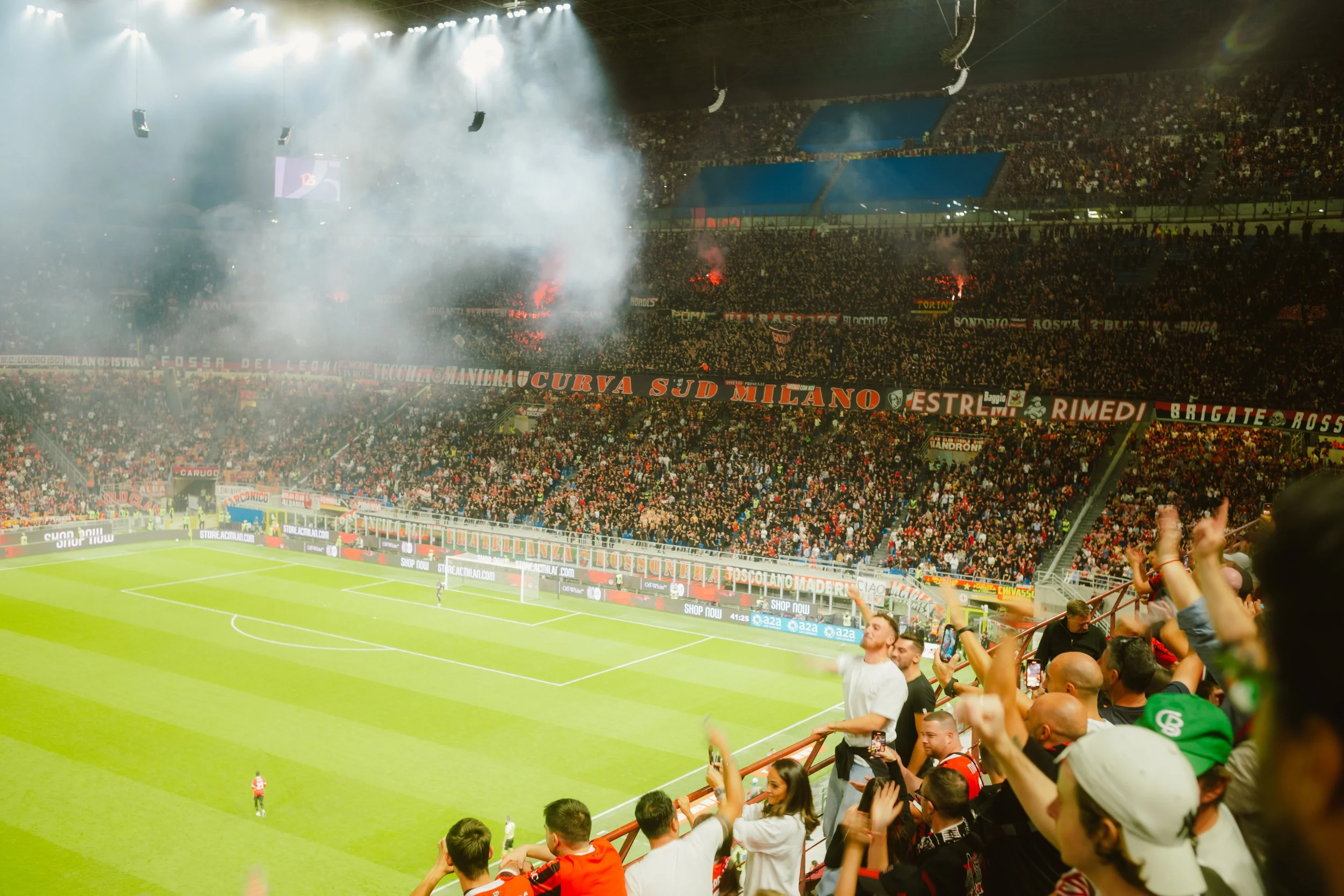 A crowded football stadium during a match with fans cheering, smoke and flares visible in the stands, and players on the field.
