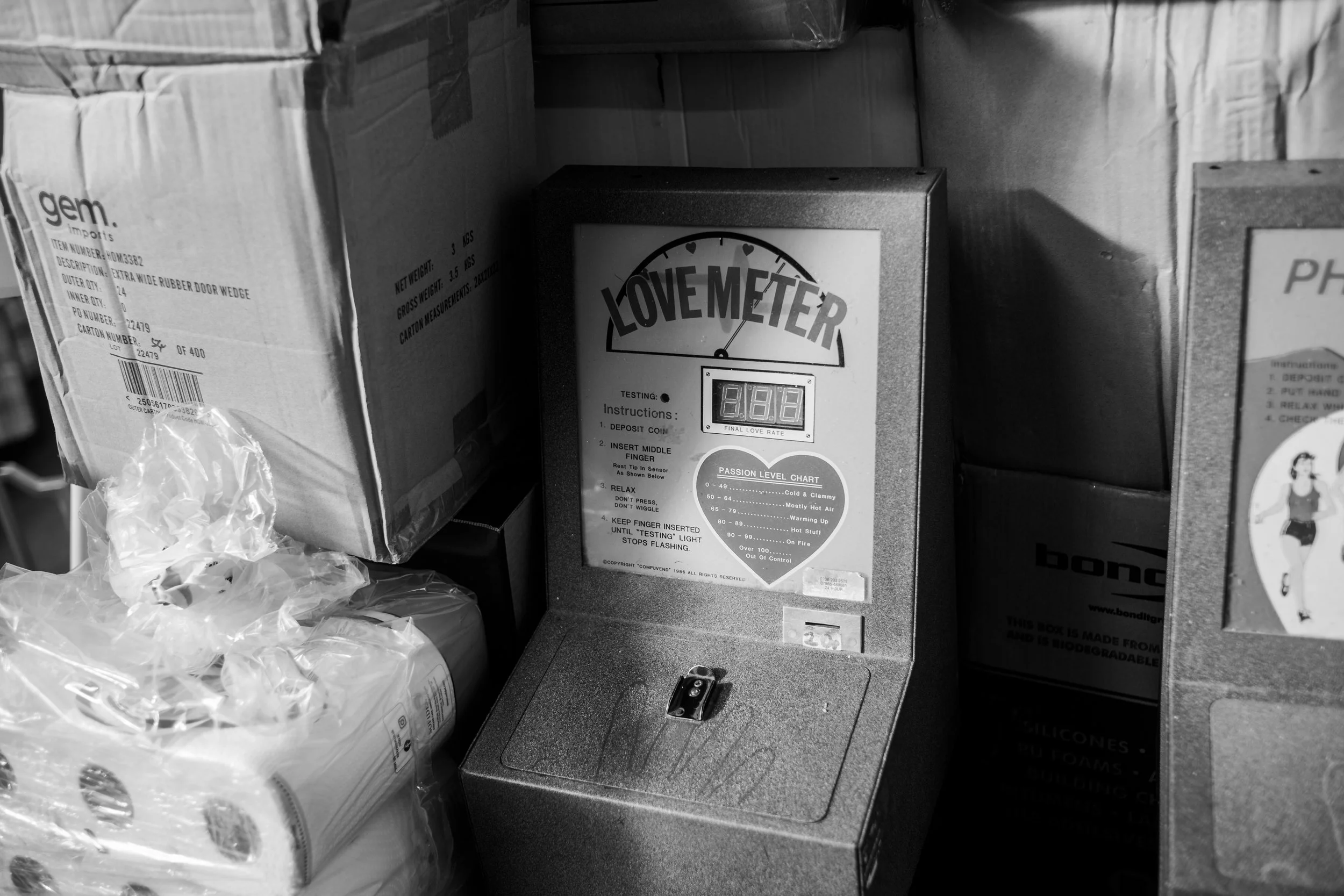 A Love Meter device is placed among cardboard boxes in a storage area. The Love Meter has a digital display and is labeled with instructions.