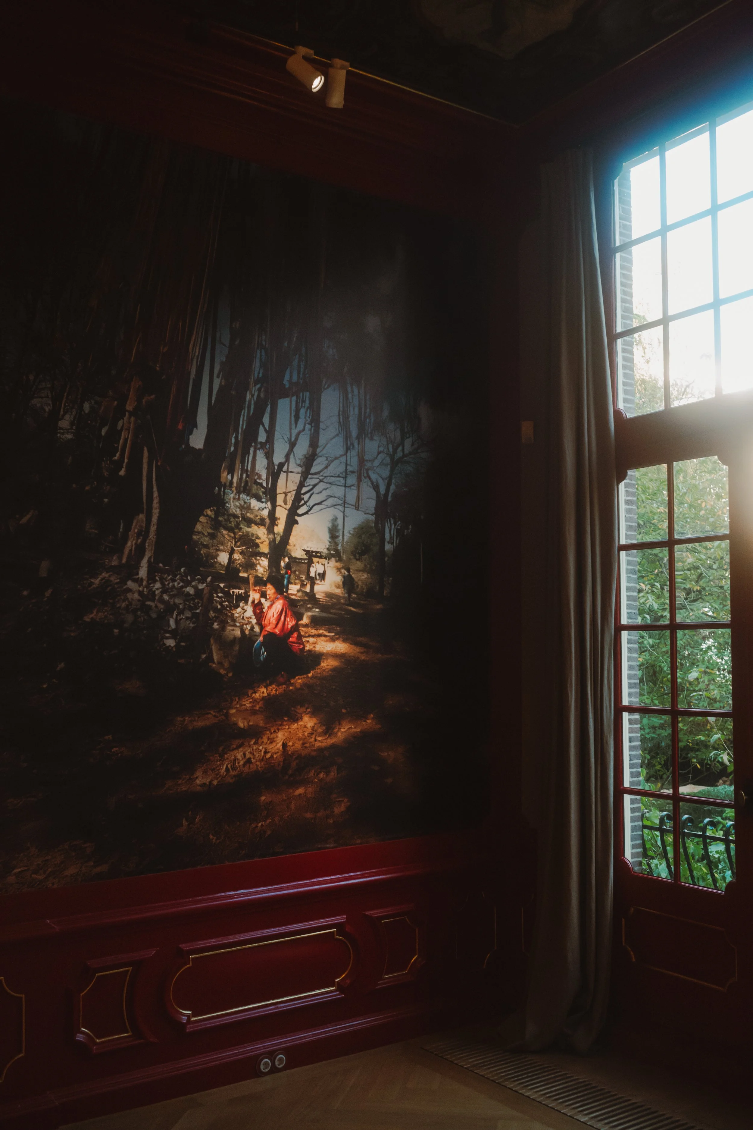 A large framed photograph of a forest scene hangs on a dark maroon wooden wall in a room, with sunlight coming through a large window with curtains on the right side.