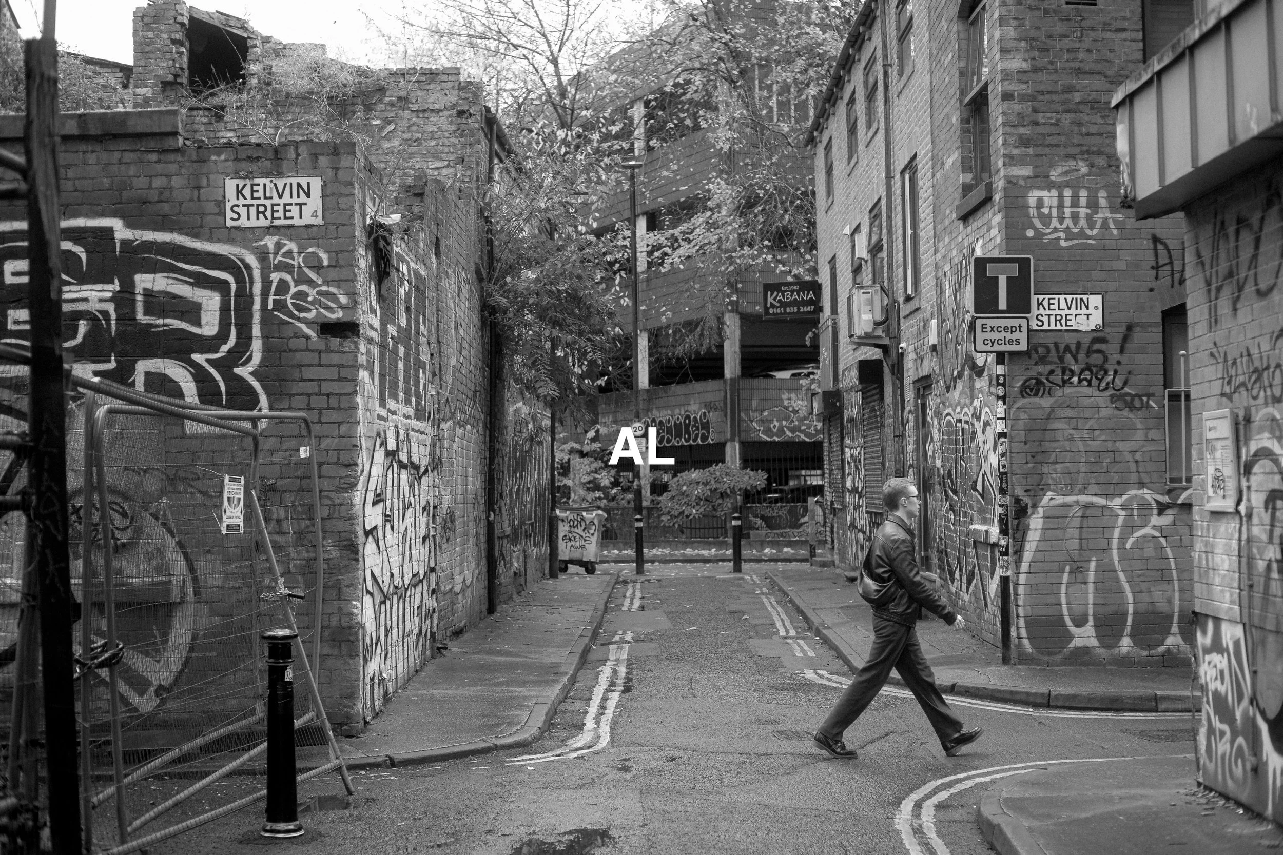 A person crossing the street at Kelvin Street, surrounded by graffiti-covered brick walls and urban buildings, black and white photo.
