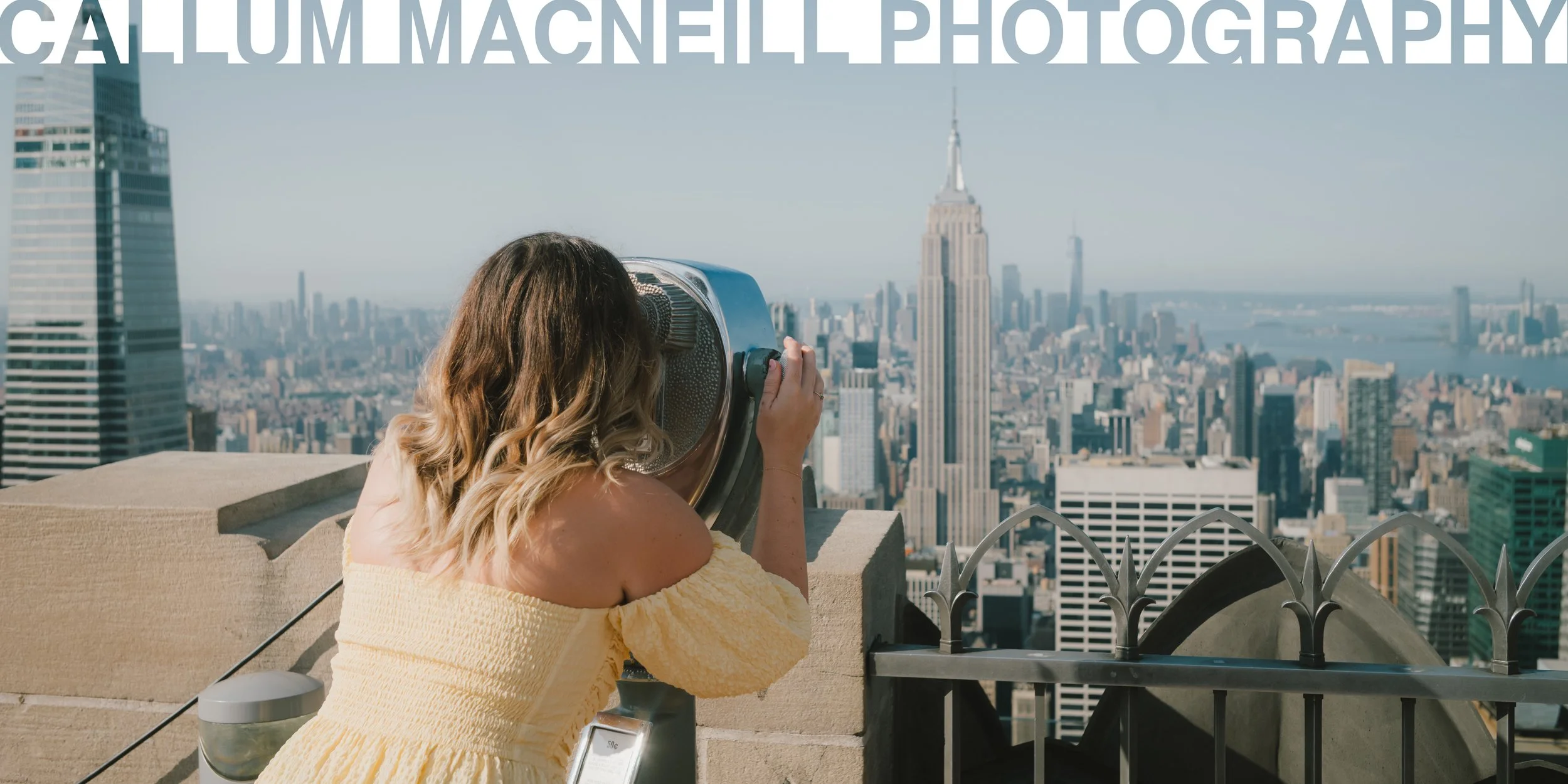 A woman with wavy blonde hair, wearing a yellow off-shoulder dress, looking through a coin-operated viewing telescope at New York City skyline, including the Empire State Building, from a rooftop observation deck.
