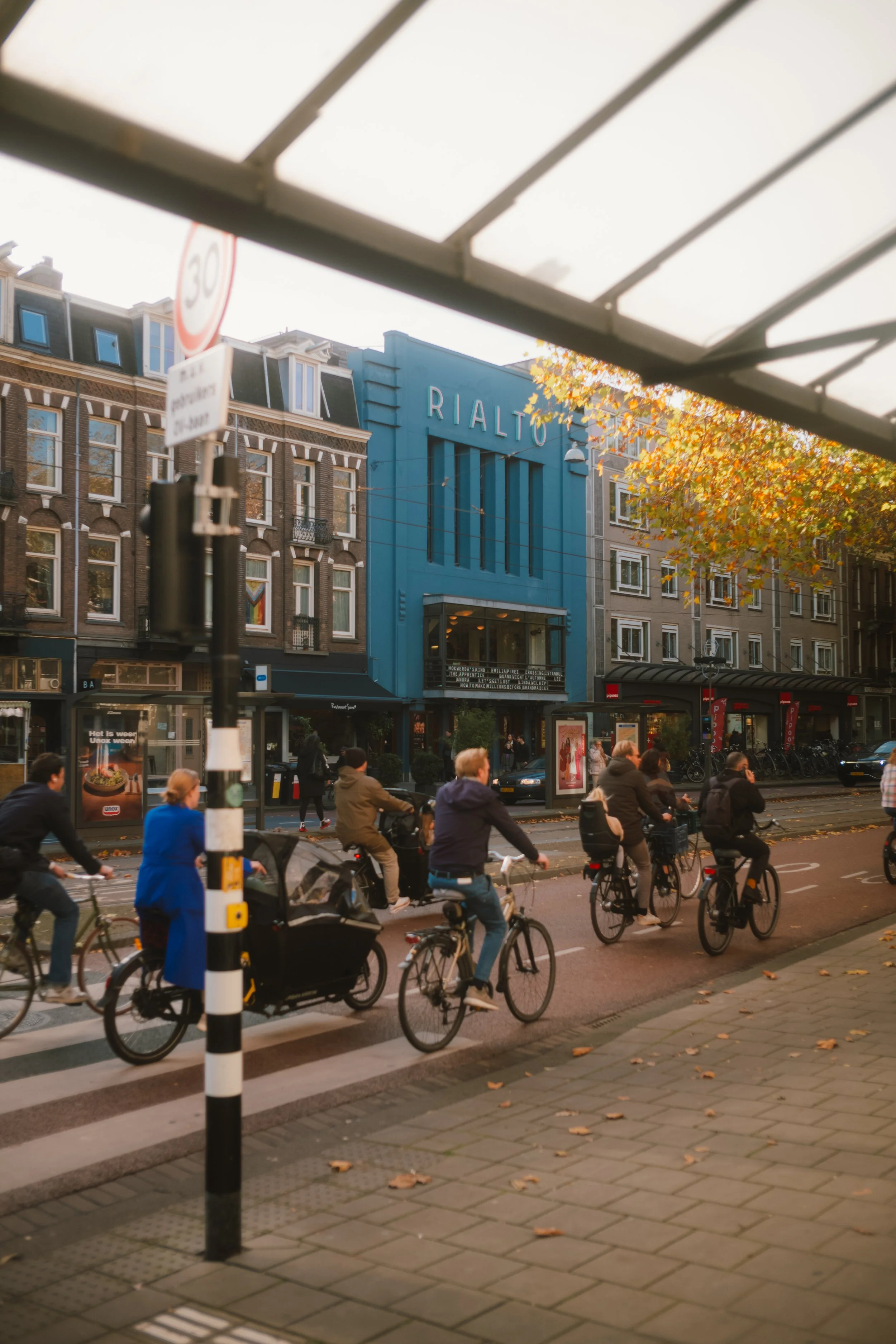 People ride bikes and walk on a city street with a blue building labeled 'RIALTO,' autumn leaves on trees, and a red and white speed limit sign.