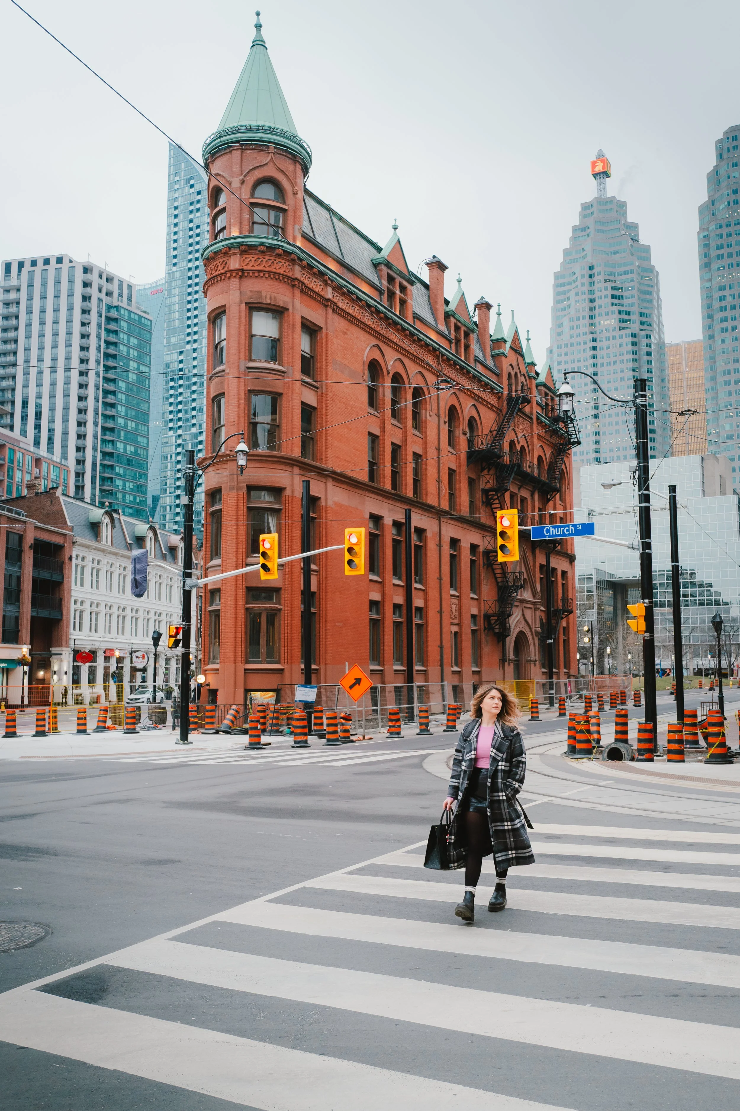 A woman walking across a crosswalk in an urban cityscape with historic red brick buildings and modern skyscrapers in the background. She is carrying a black bag, wearing a plaid coat, black pants, and boots.