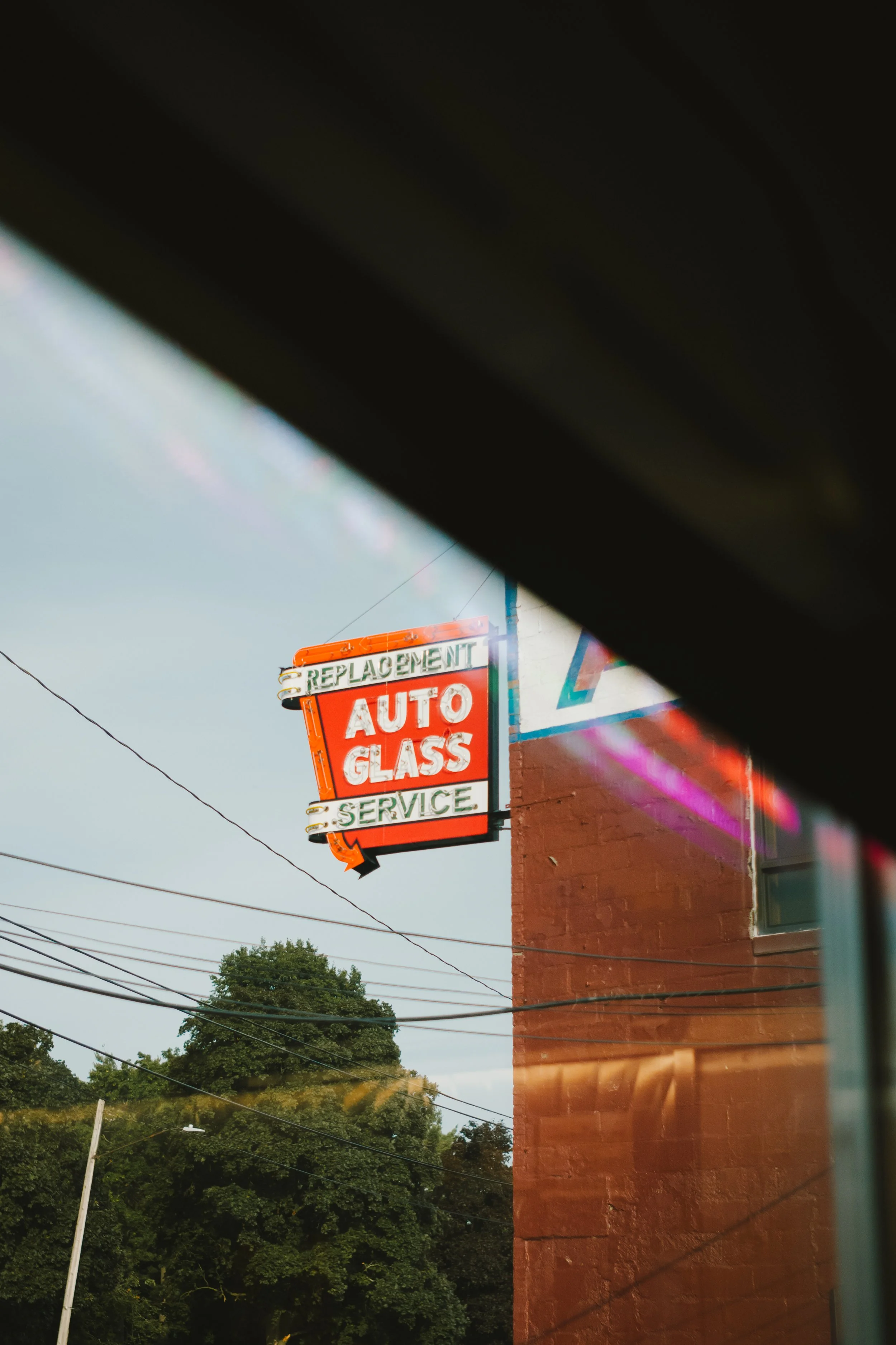 A neon sign that reads "Replacement Auto Glass Service" on a building next to a smaller sign for auto glass