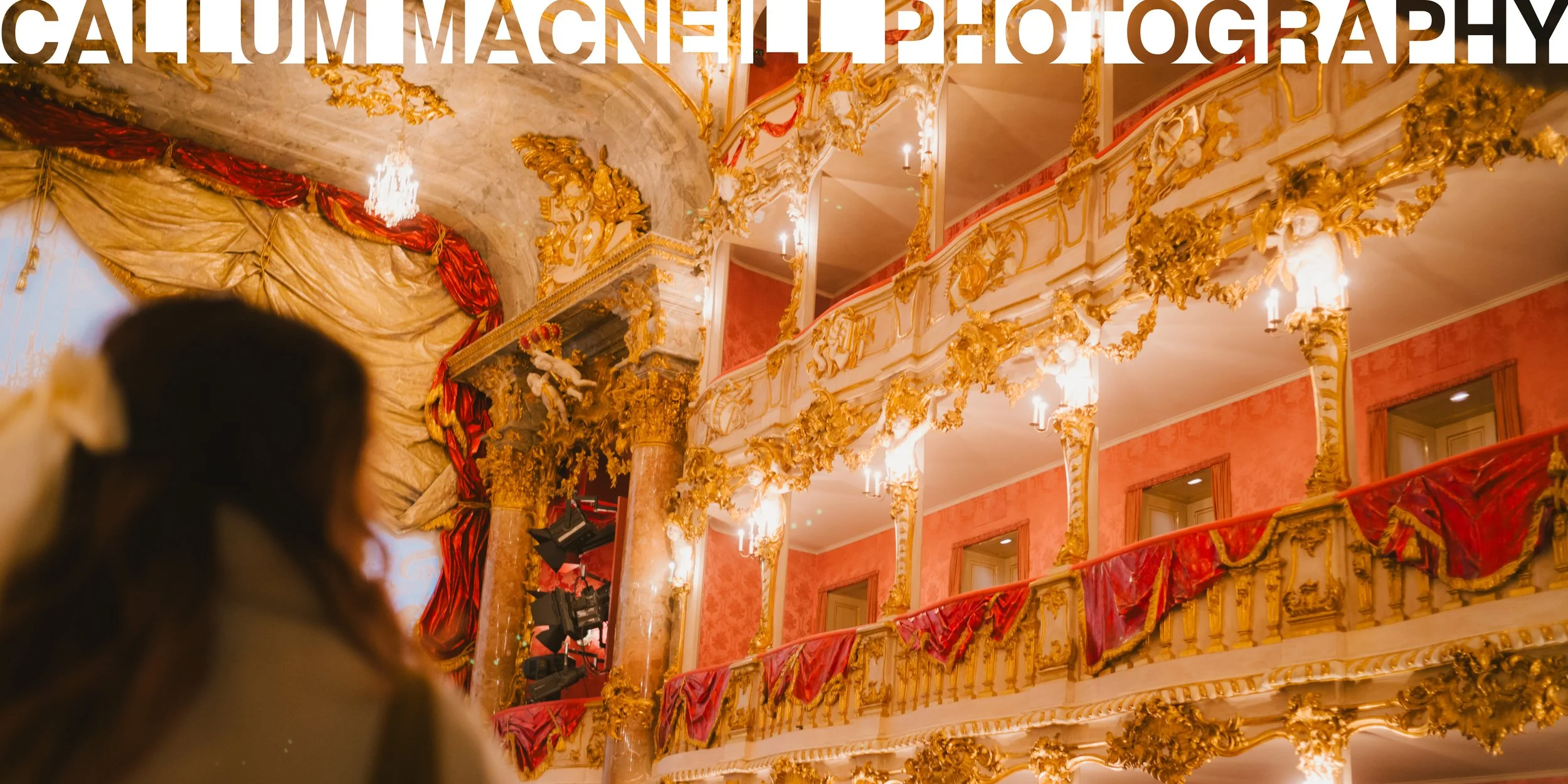 Interior of an opulent theater or opera house with ornate gold decoration, red velvet curtains, and balconies adorned with gilded sculptures and intricate details.