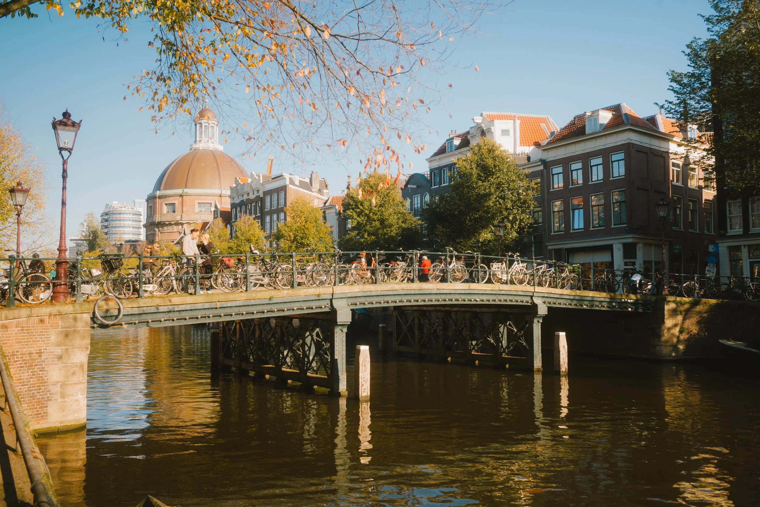 A cityscape featuring a river with a bridge, bicycles parked along the riverbank, historic buildings with red-tiled roofs, a domed building, and trees with autumn leaves under a clear blue sky.