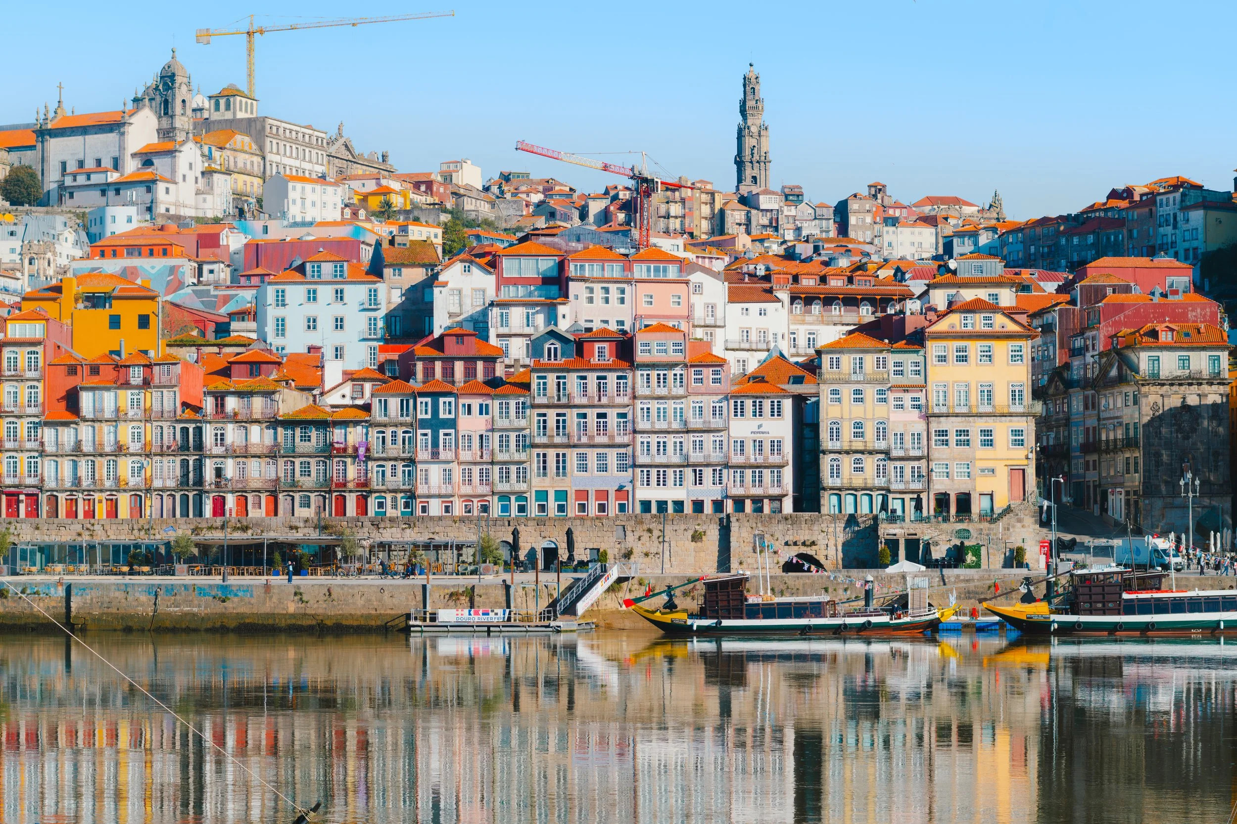 Colorful buildings with red roofs along a waterfront under a clear blue sky in Porto, Portugal. A tower and construction cranes are visible on a hill in the background.