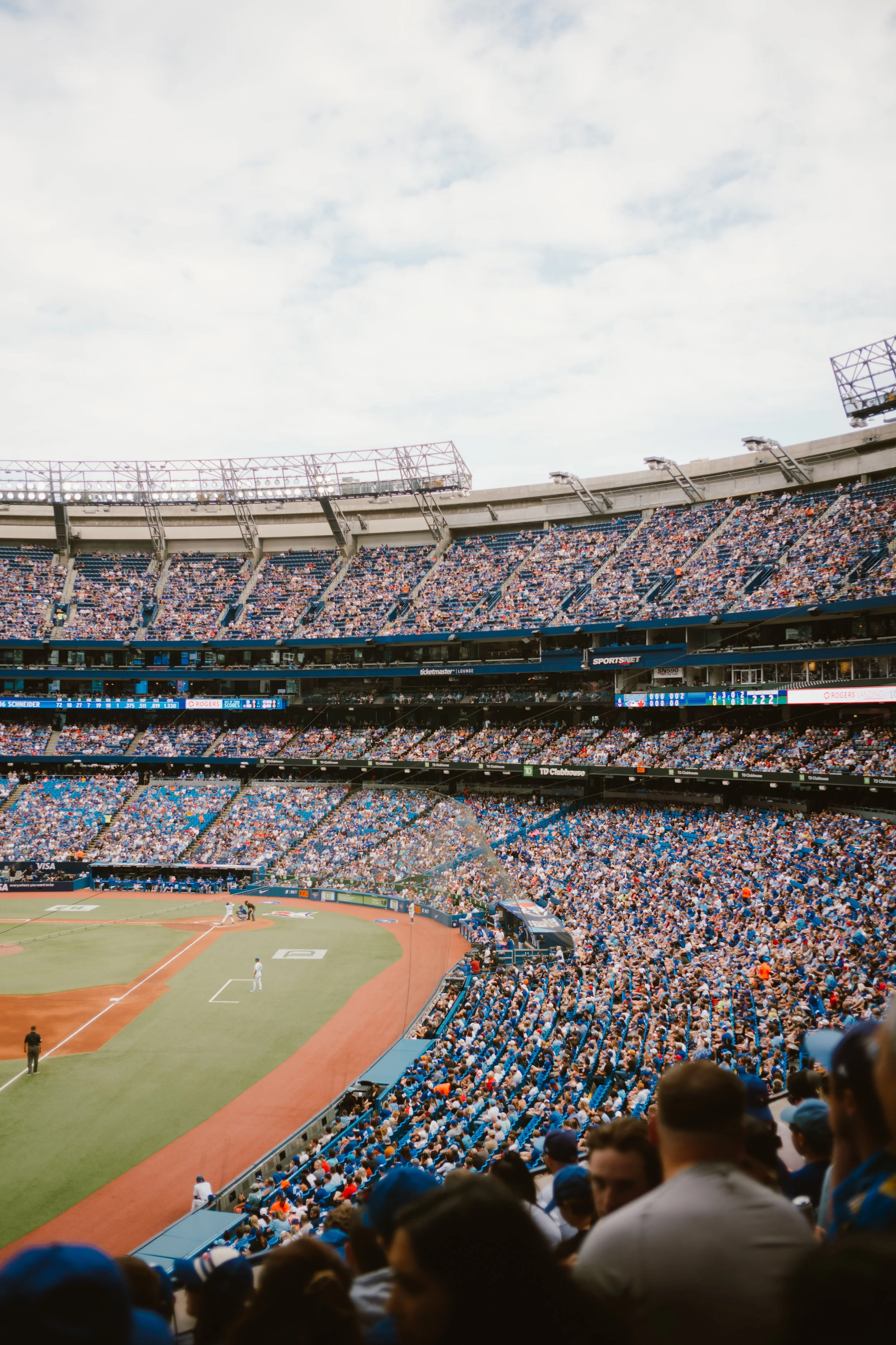 A large baseball stadium filled with spectators, with players on the field preparing for the game under a partly cloudy sky.