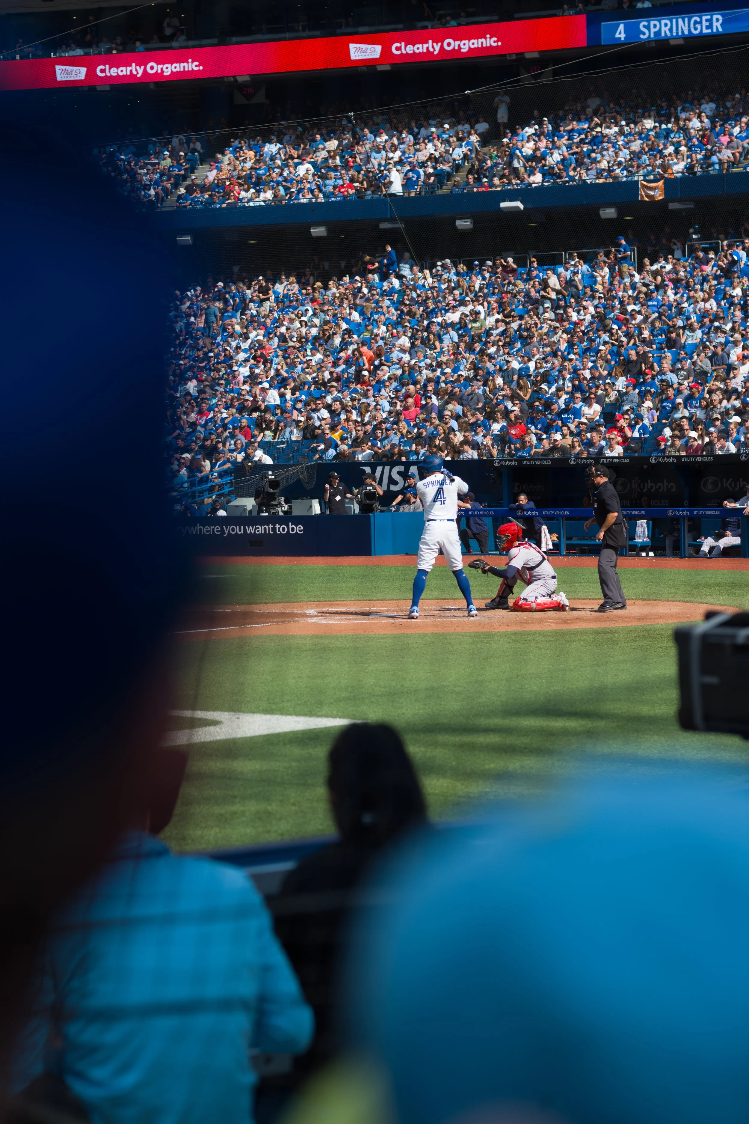 View of a baseball stadium's crowd from behind the dugout, with a batter preparing to hit and a catcher and umpire behind home plate, and fans filling the stands.