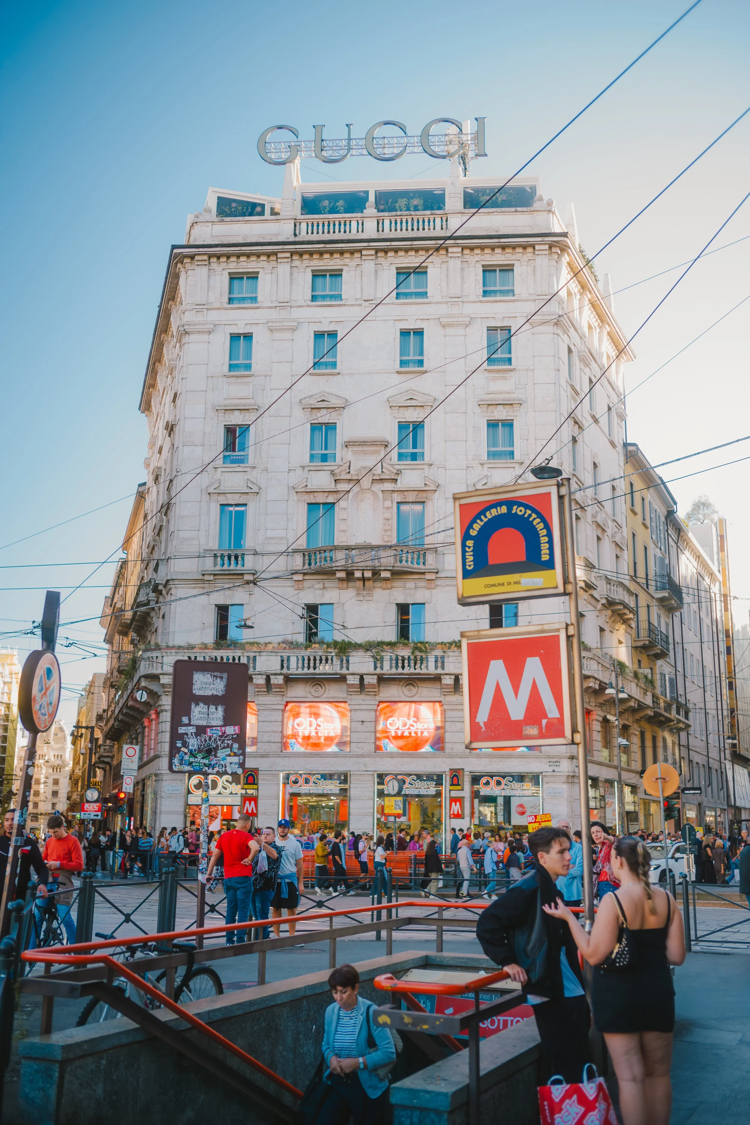 A busy street scene in a city with a tall building that has a large Gucci sign on the roof. People are walking on the sidewalk, and there are signs for a metro station and stores. The sky is clear and the sun is shining.
