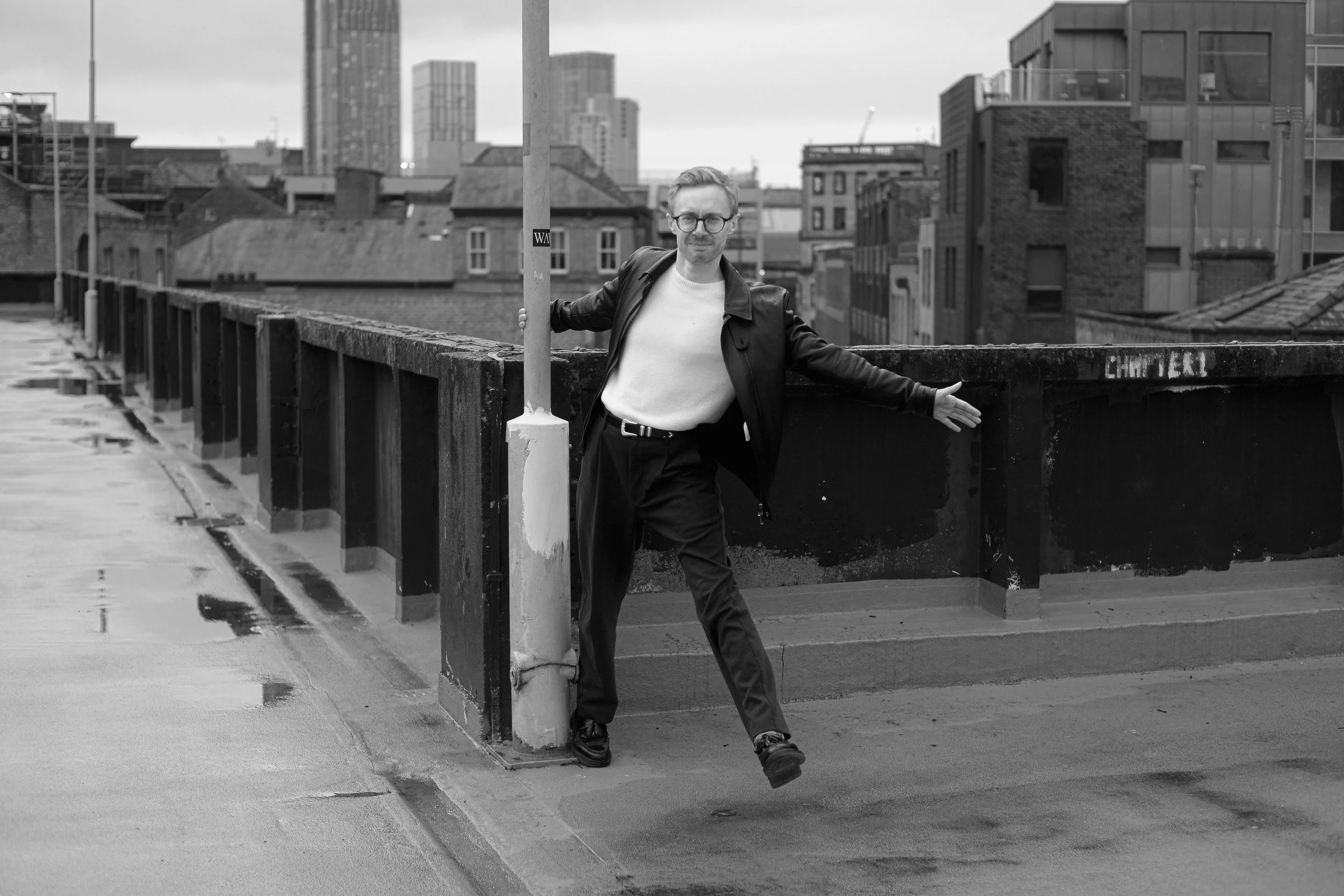 A man with glasses, wearing a white shirt and black jacket, standing on a city street. He is leaning on a concrete barrier with his left arm, with an urban skyline of high-rise buildings in the background. The photo is in black and white.