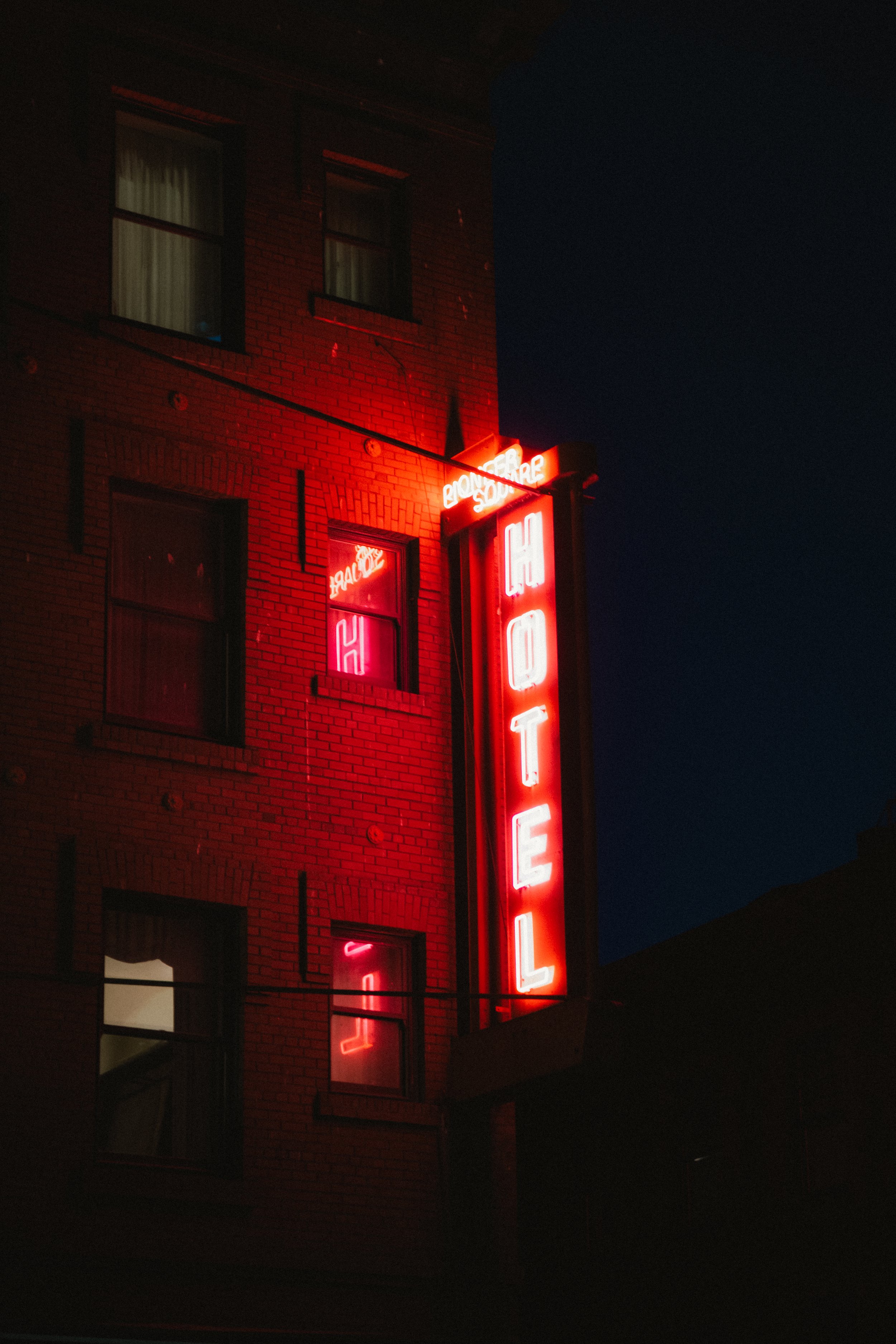 Night view of a brick building with a vertical red neon sign reading 'HOTEL' and a small illuminated sign with the word 'BALCONY' near the top, against a dark sky.