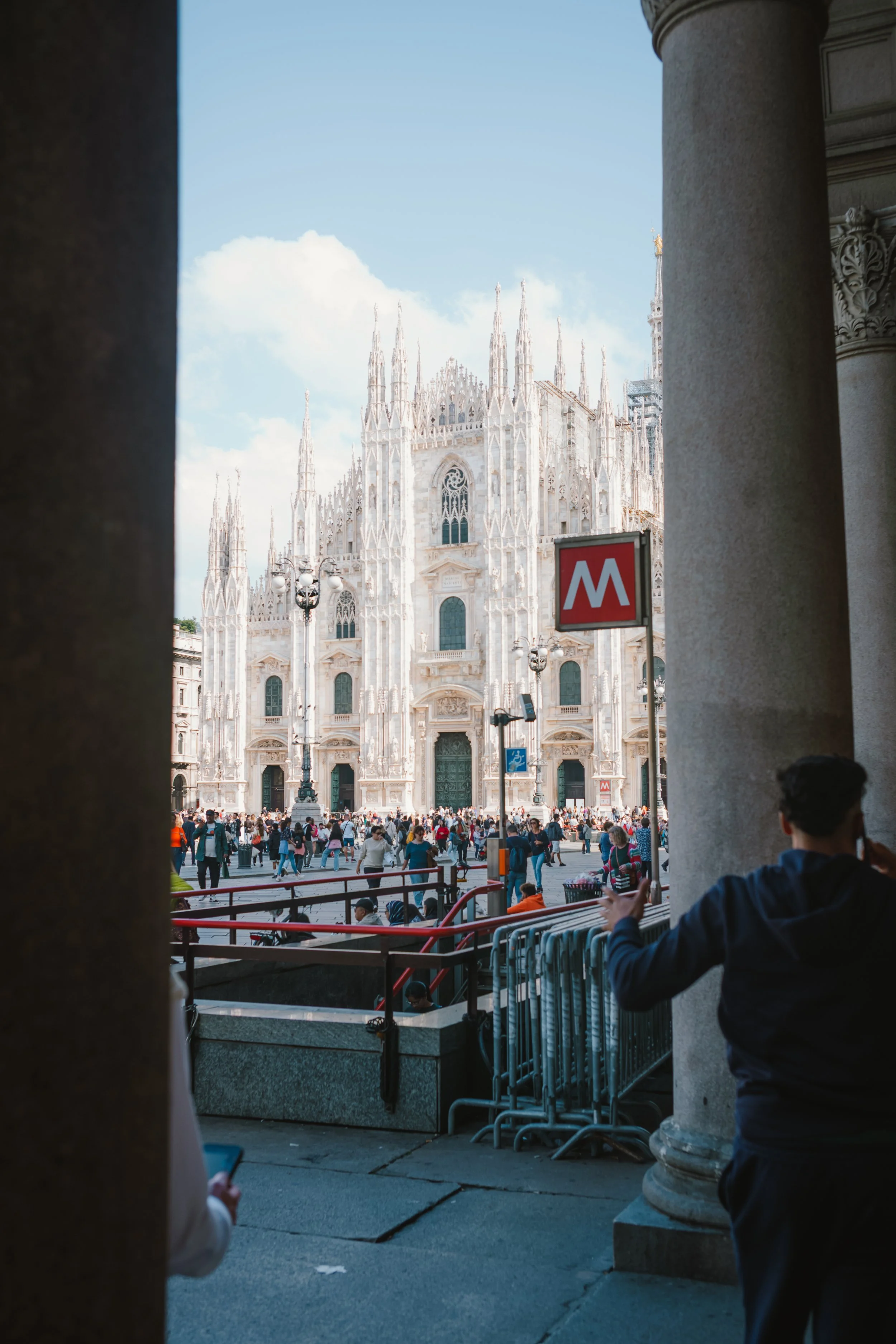 View of a large cathedral with elaborate Gothic architecture, seen from behind columns and a crowd of people in front, as seen from a nearby underground metro station entrance. The scene includes a red and white metro sign and bright daylight.