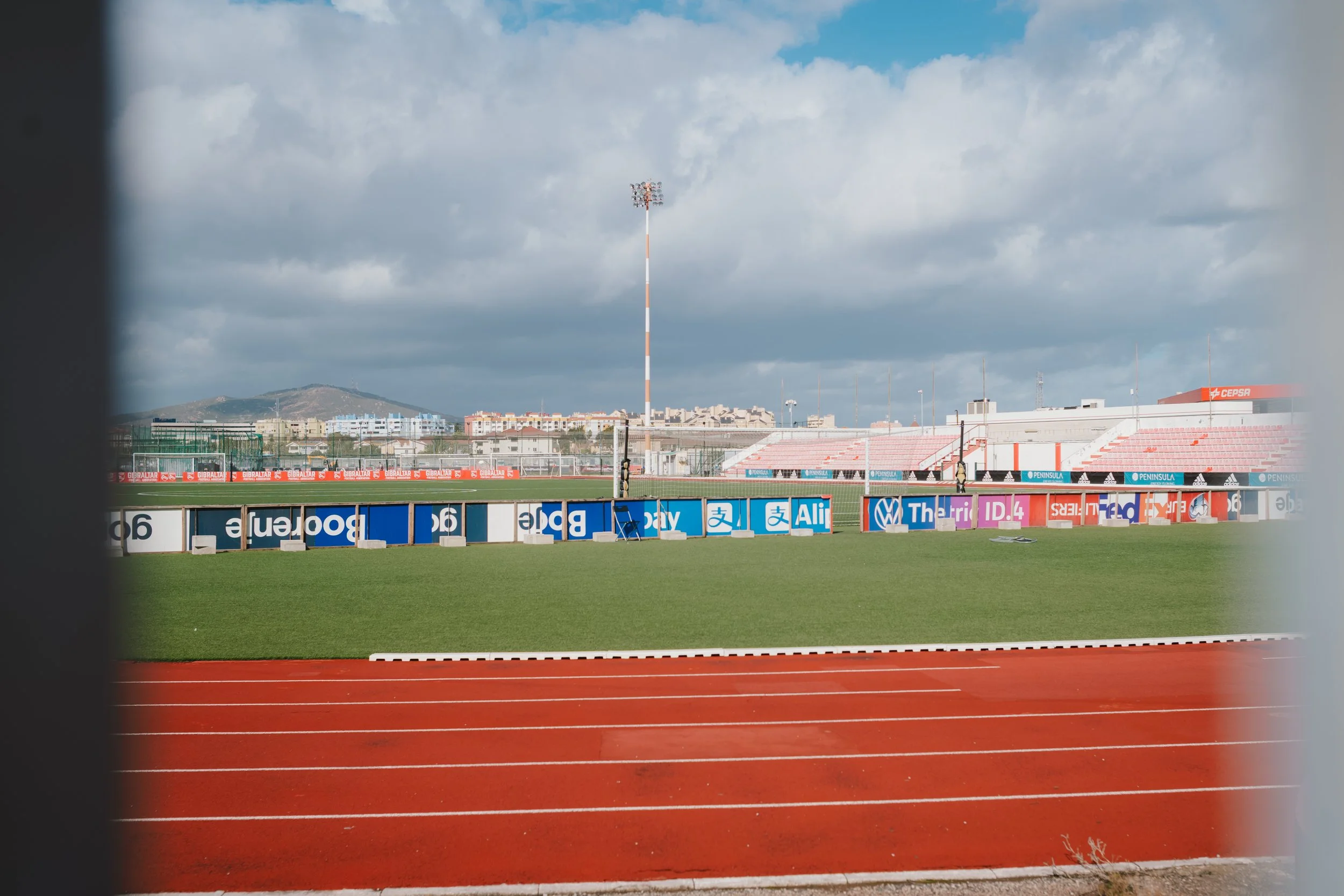 View of a sports stadium with a running track, green field, advertising banners, and empty seating stands, under a partly cloudy sky.