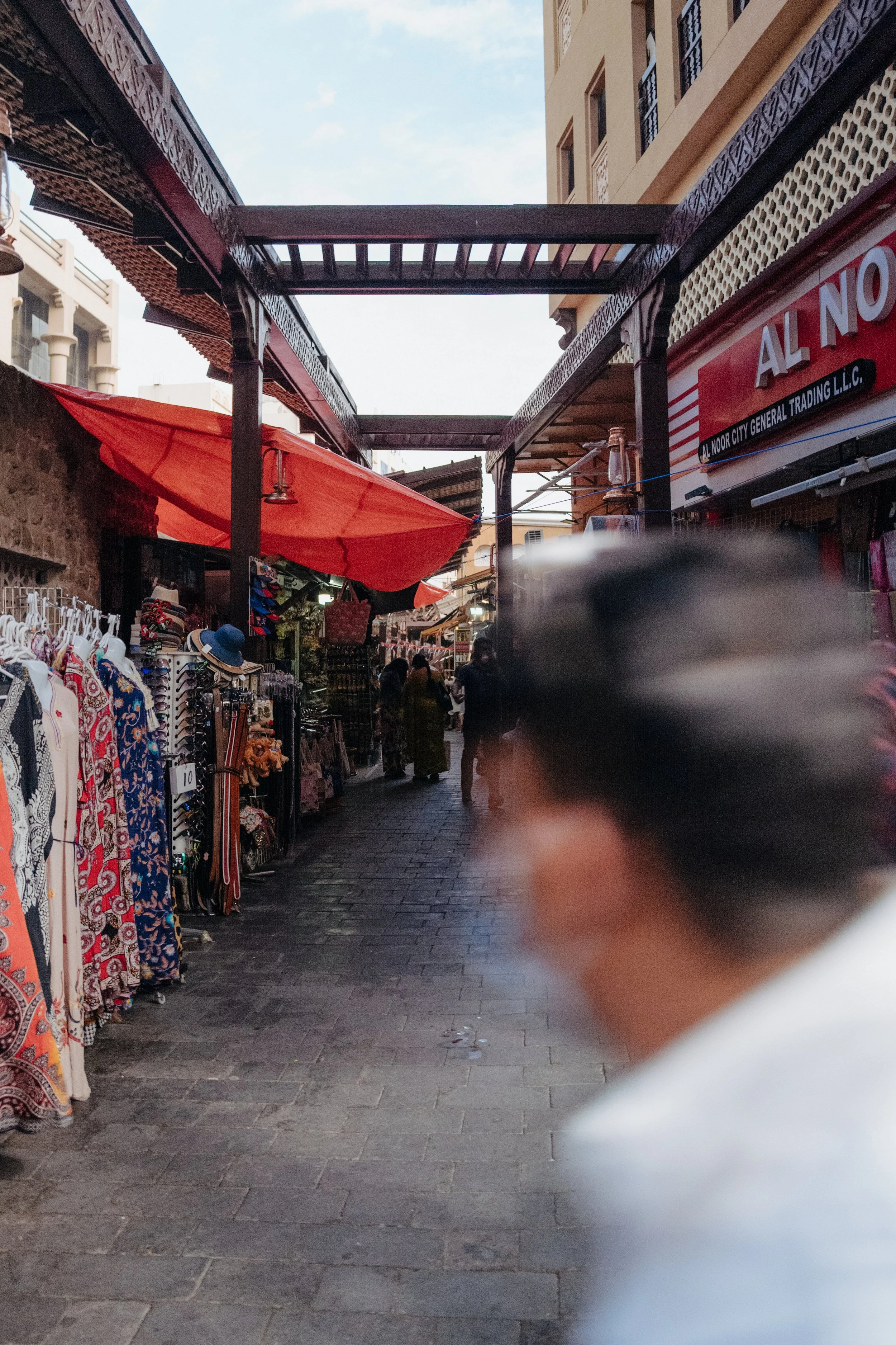 A crowded outdoor marketplace with clothing and accessories on display, shops on either side, and a few people walking along the cobblestone street. Overhead, there are buildings with balconies and a partly cloudy sky.