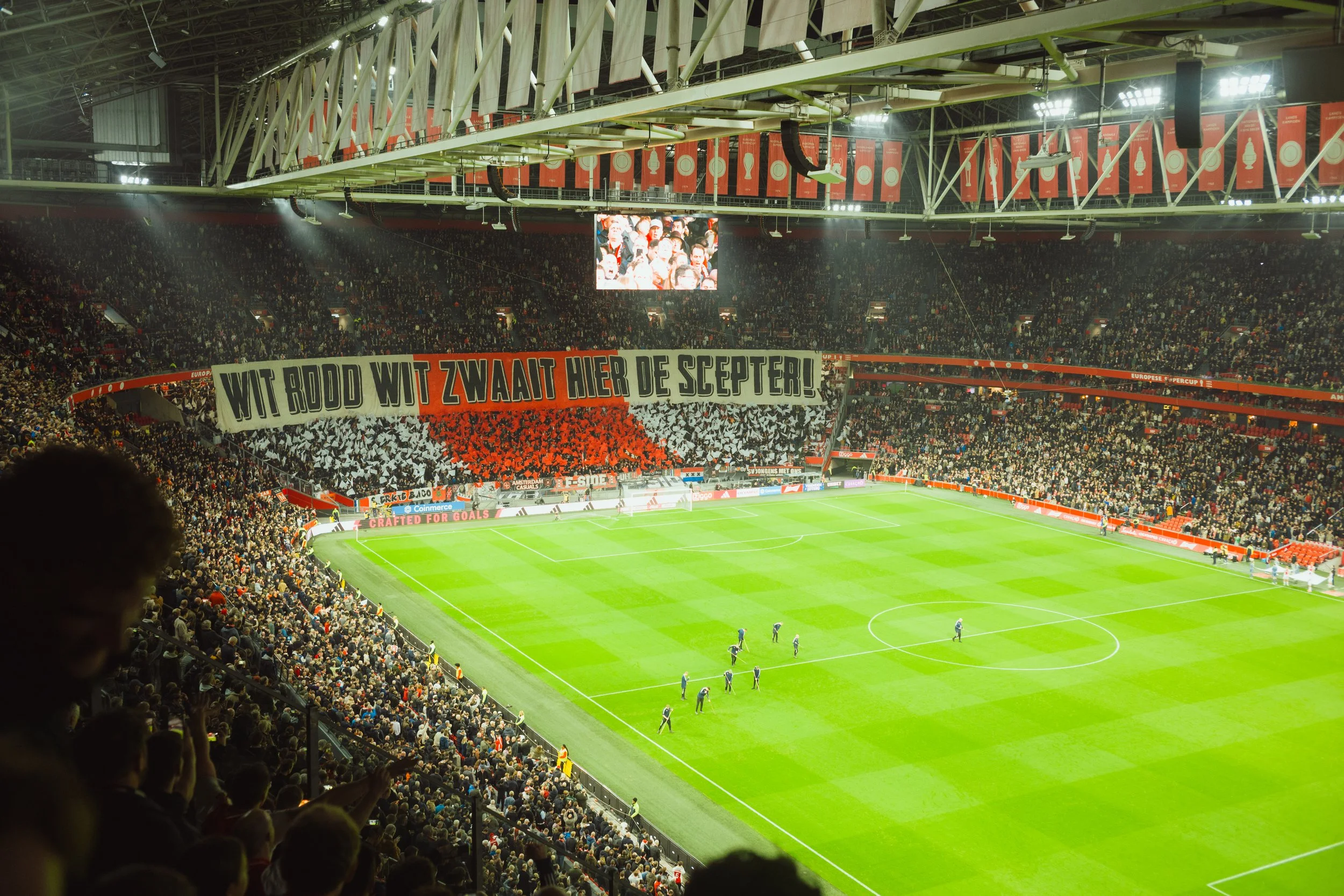 A crowded soccer stadium with fans in the stands, a large banner with Dutch text, and a giant screen displaying fans.