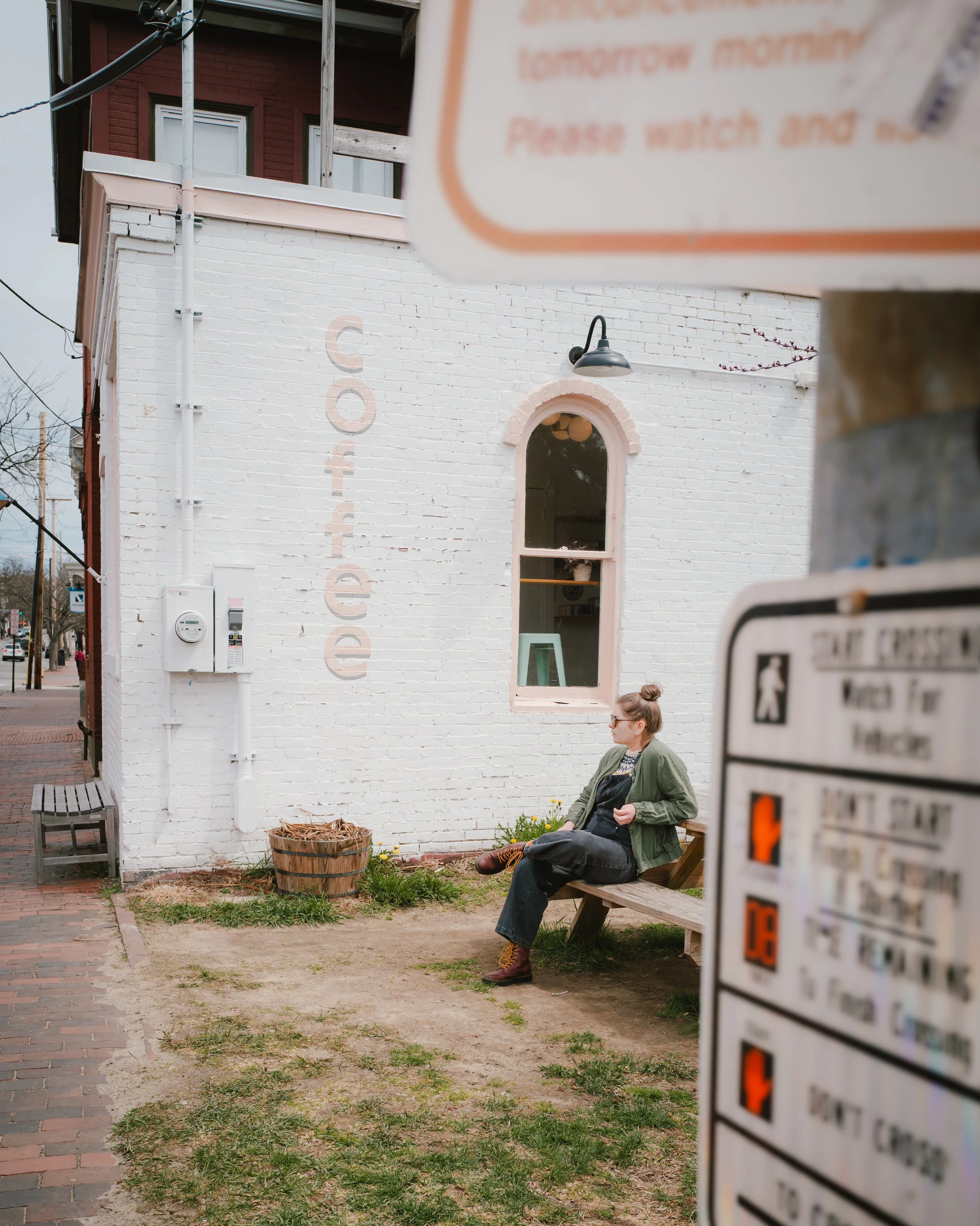 A woman with glasses, a bun hairstyle, and a green jacket sitting on a wooden bench outside a white brick building. The building has a sign with text that is partially obscured. There is a potted plant with yellow flowers near the woman. In the foreg