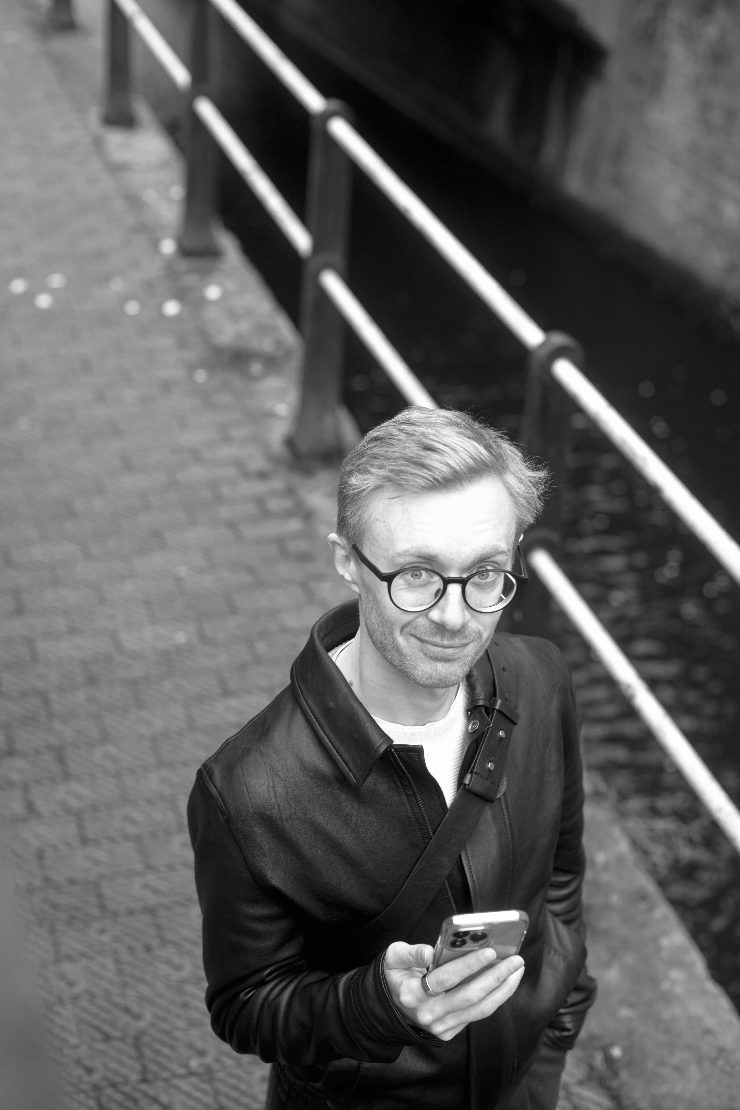 A man with glasses and short hair, wearing a black leather jacket, is looking up at the camera while holding a smartphone in his right hand. He is standing outdoors near a bridge or railing.