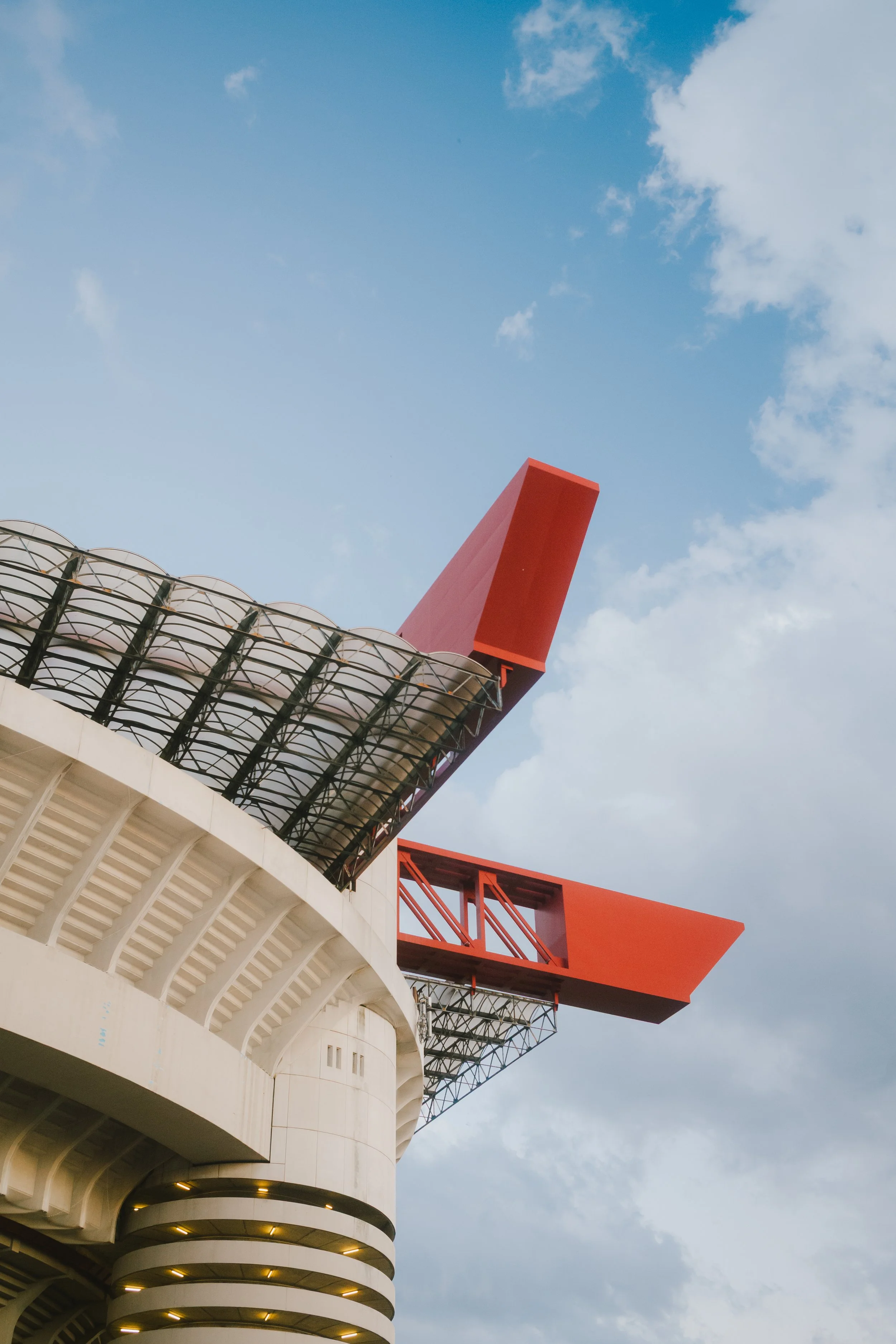 Exterior view of a stadium with a white arched facade and a rooftop structure with a bright orange beam extending outward, against a partly cloudy sky.