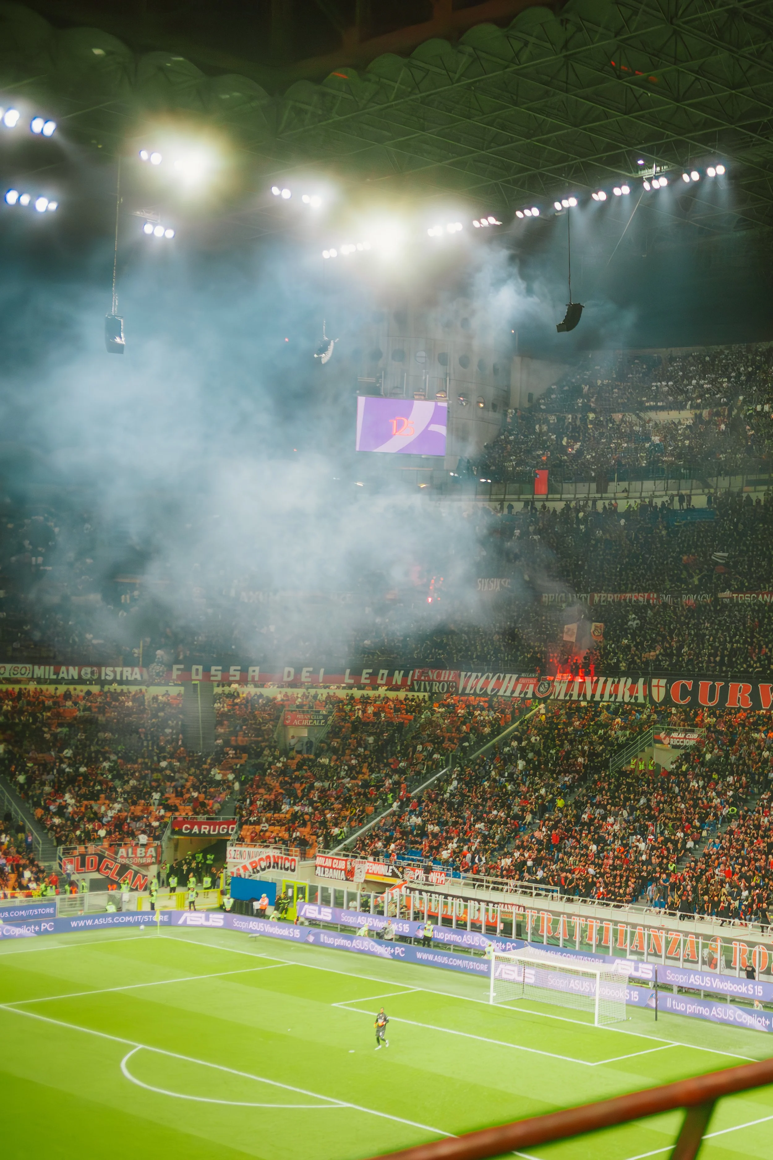 A soccer stadium filled with spectators, some waving flags. Bright stadium lights illuminate the field, and a player is on the field seen near the corner kick area. Smoke or fog effects are visible in the stands, creating a dramatic atmosphere.