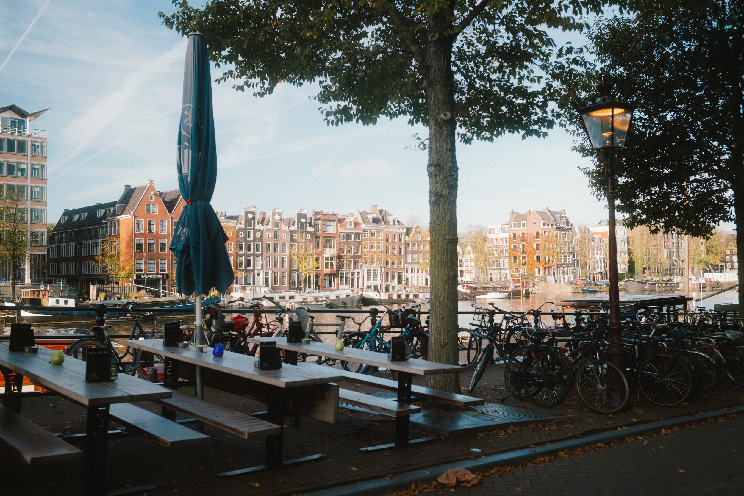 Outdoor seating area with wooden picnic tables and benches, bicycles parked nearby, a large tree providing shade, a lamppost, and a canal with boats in the background. Several buildings and houses line the canal, under a partly cloudy sky.