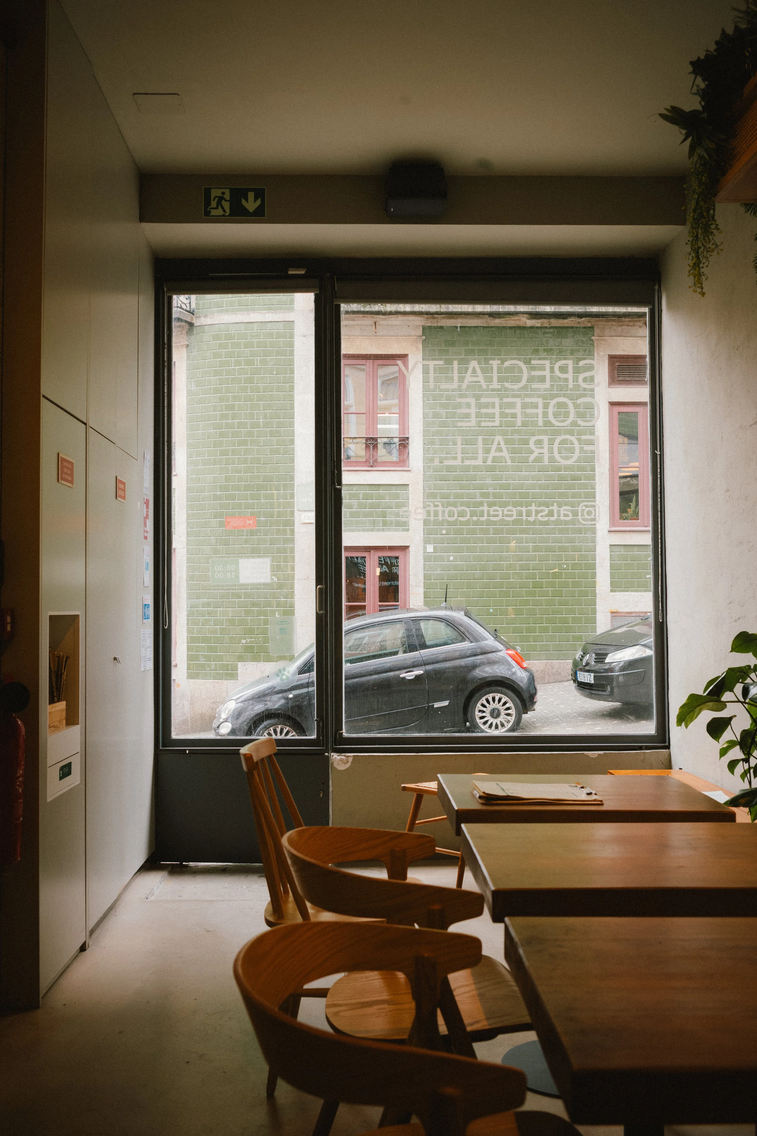 Interior view of a cafe with wooden chairs and tables near a window showing green building facades and parked cars outside.