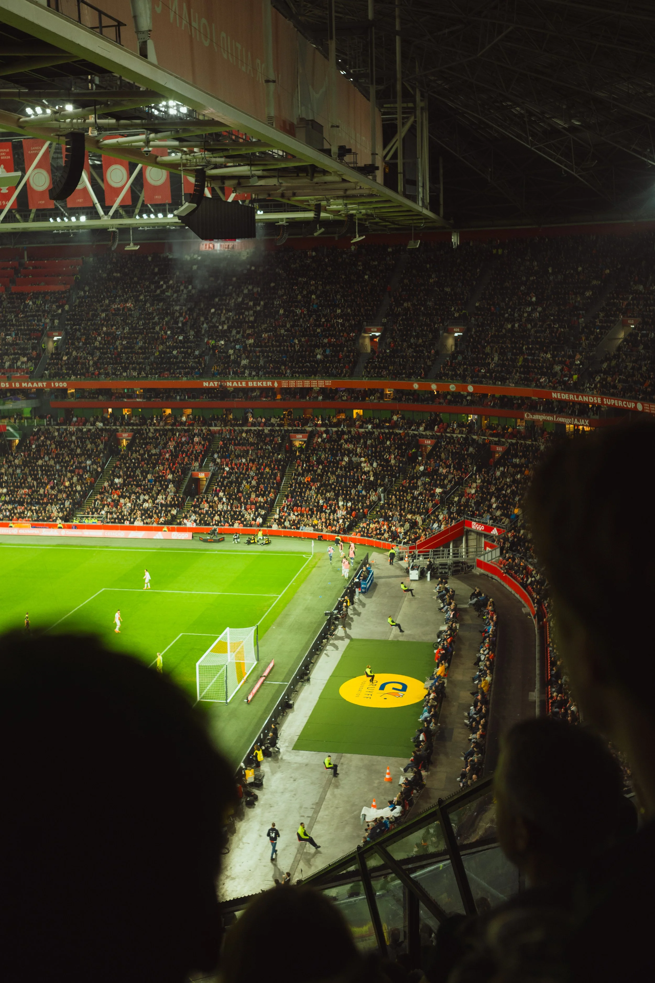 View of a soccer stadium filled with spectators, taken from behind a protective barrier with the field visible in the foreground, and players on the field.