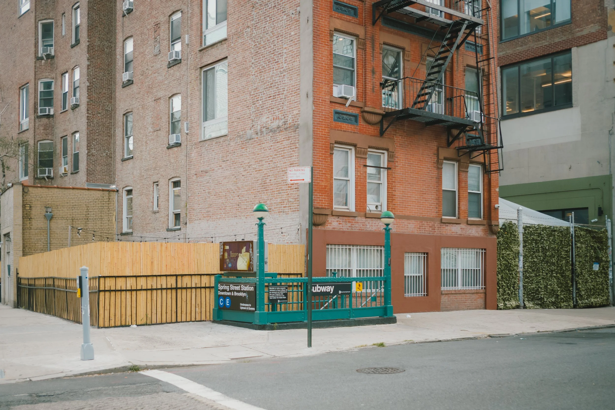 Entrance to Spring Street Station subway with green signage for C and E lines, surrounded by a wooden fence and urban buildings.