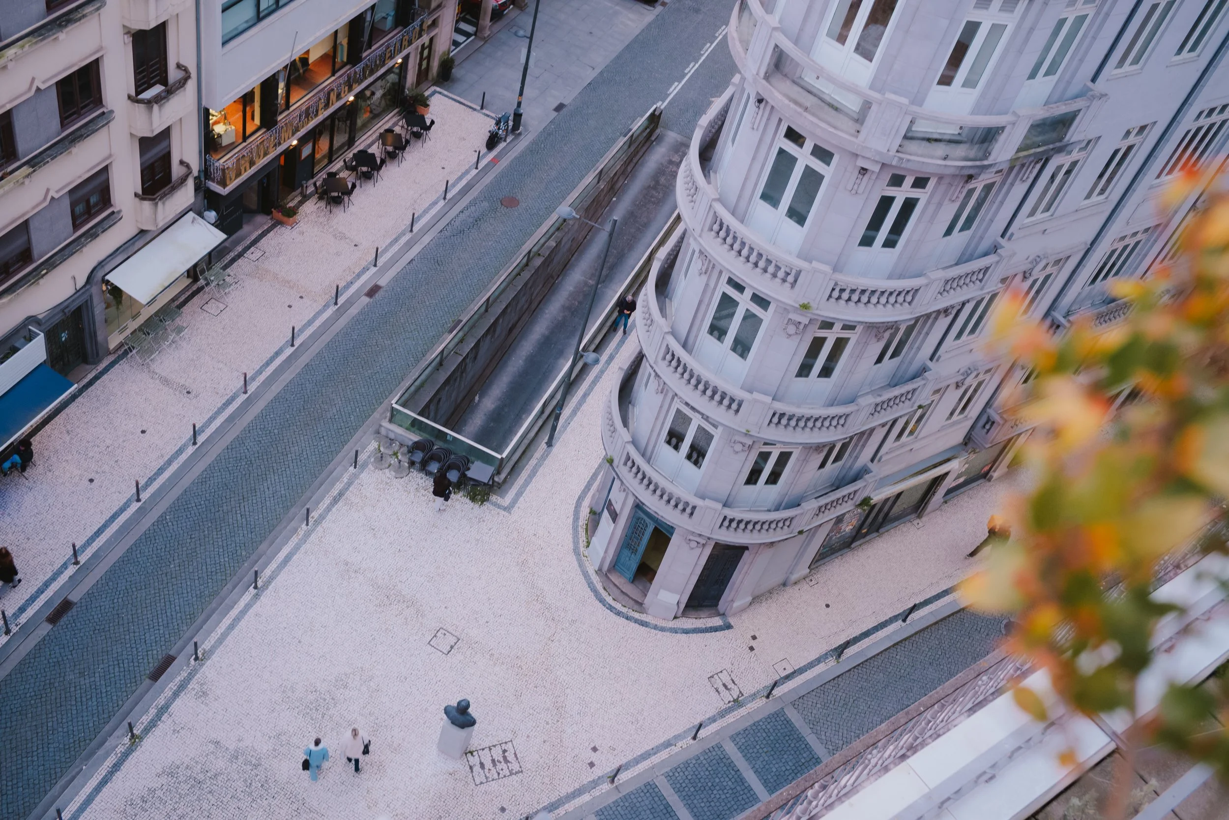 View of a city street from a high angle showing a curved white building with multiple windows and balconies, a sidewalk with outdoor seating, street lamps, and a few people walking.