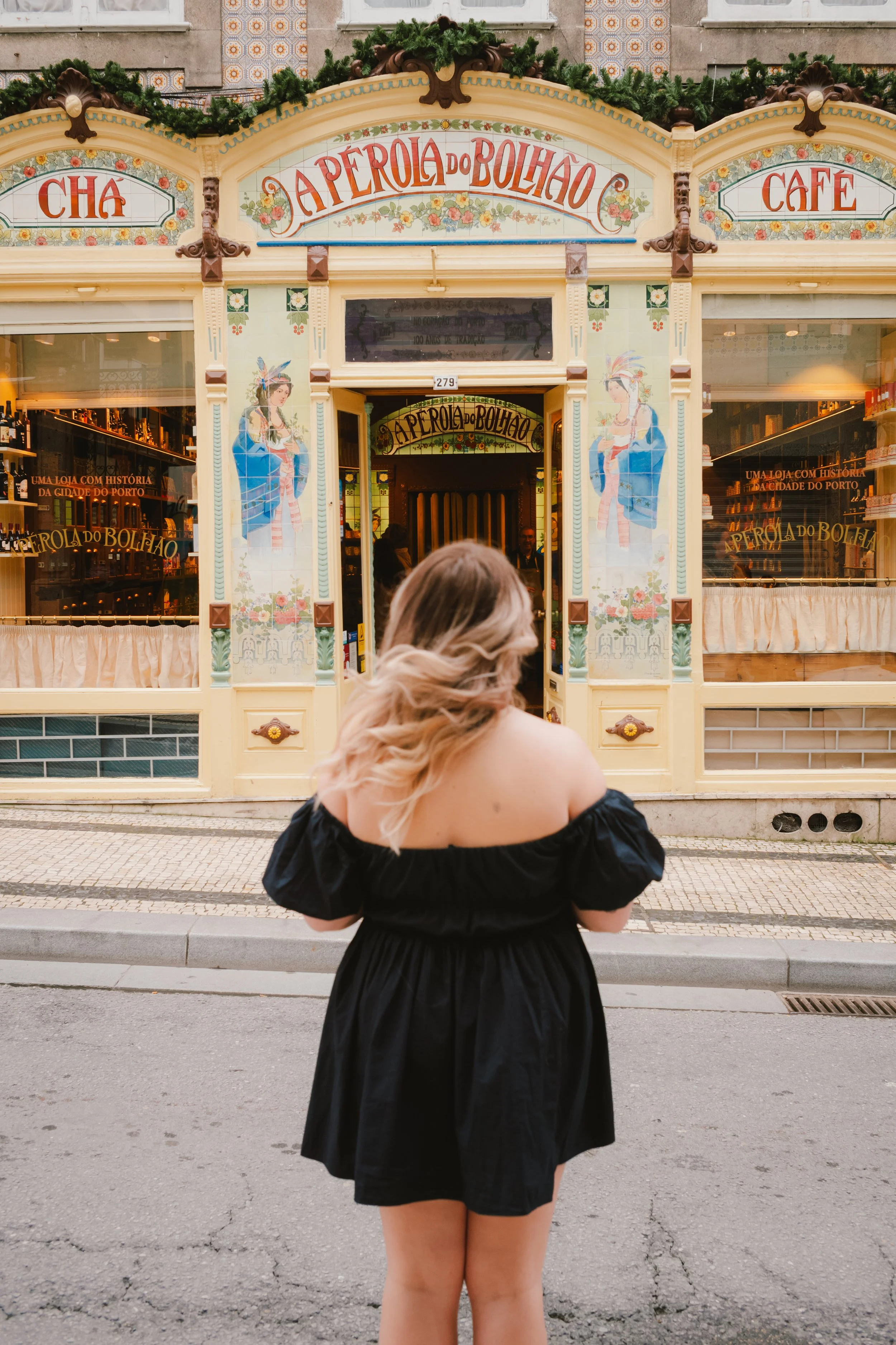 A woman wearing a black off-the-shoulder dress standing in front of a decorative storefront with colorful illustrations and signs for a tea shop, cafe, and bakery. The woman has long, wavy blonde hair and is facing away from the camera.