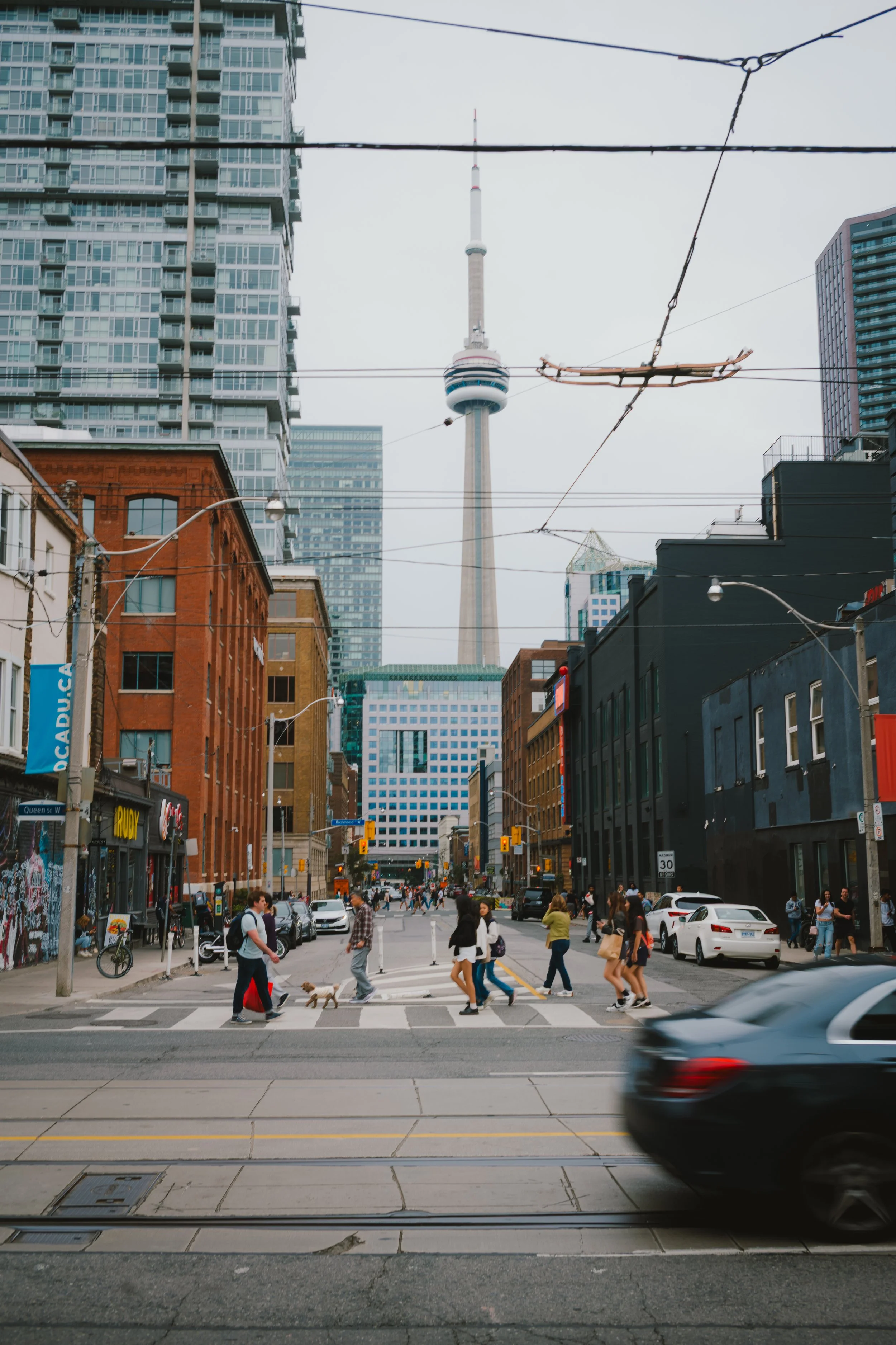 Street scene in downtown Toronto with pedestrians crossing at a crosswalk, cars on the road, and the CN Tower visible in the background amidst tall buildings.