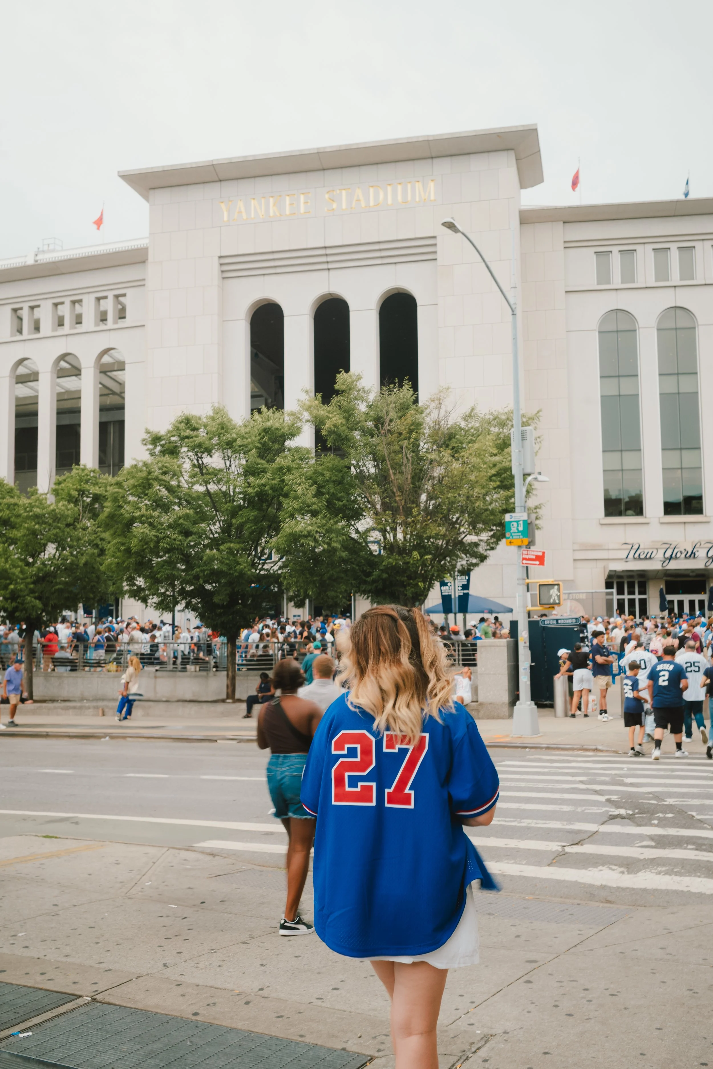 People gather outside the Yankee Stadium in New York City, with some wearing New York Giants football jerseys, including a woman in a blue jersey with the number 27.