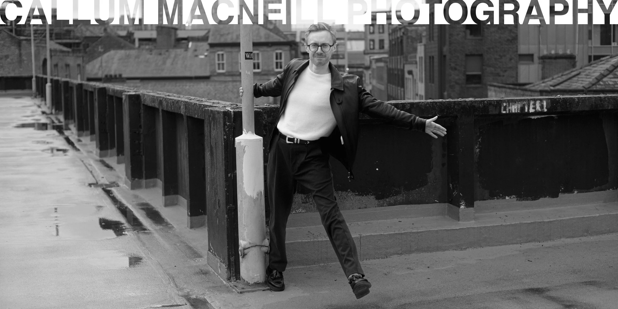 A man wearing glasses, a white shirt, and a leather jacket standing on a rooftop with a cityscape in the background. He is holding a pole and gesturing with his other hand.