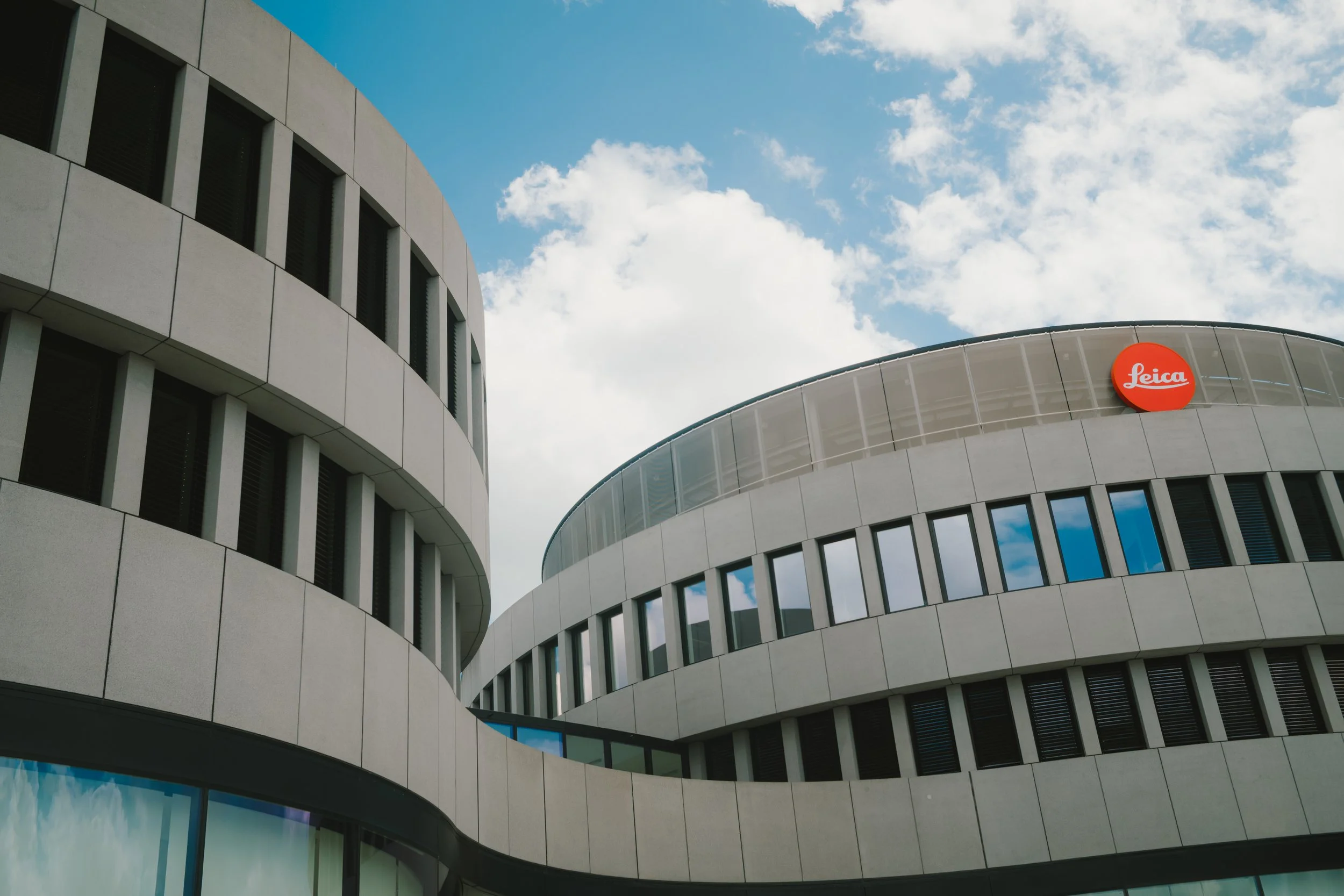 Modern building with curved architecture and large windows, featuring a red Leica logo on the facade, set against a blue sky with scattered clouds.