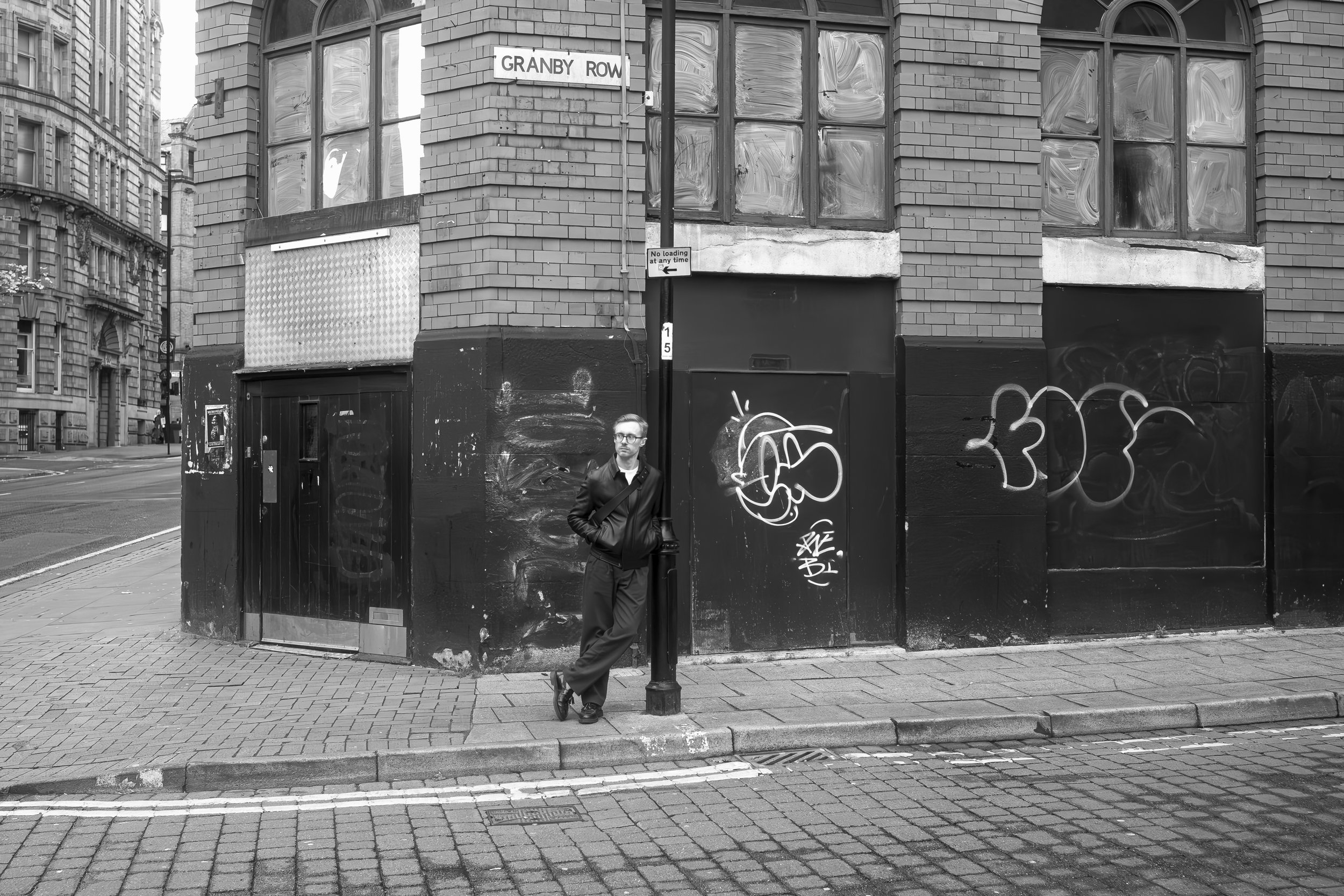 A man leaning against a streetlamp on a city sidewalk, with a brick building behind him covered in graffiti, and street signs reading 'Granby Row' and 'No loading at any time.'