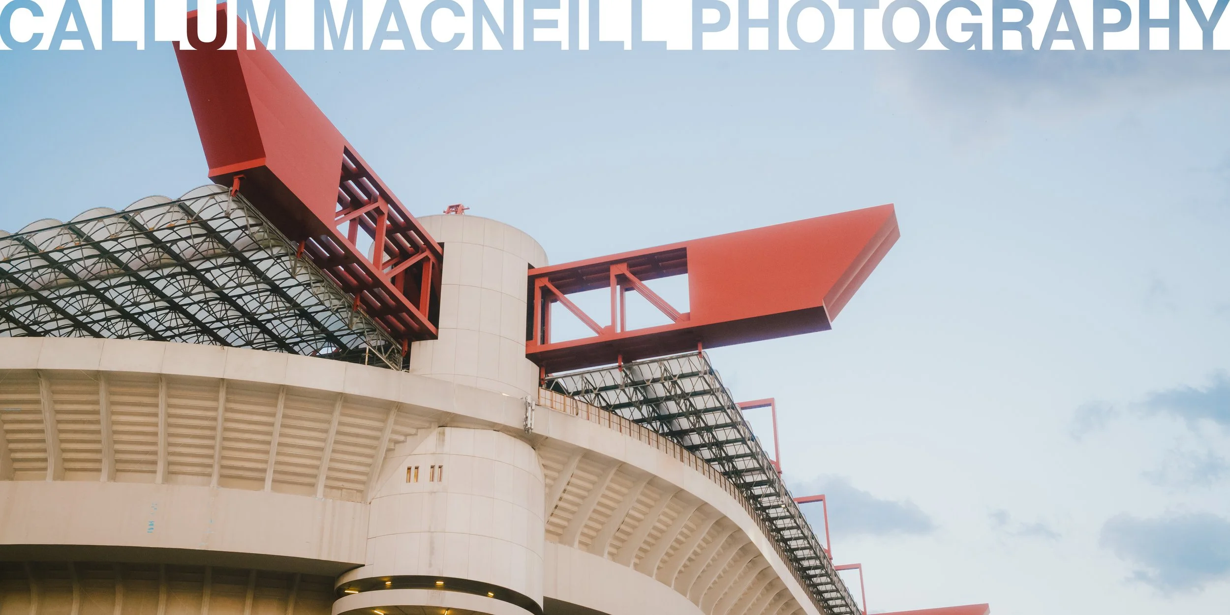 Close-up of the upper section of a stadium with red structural elements and a modern architectural design against a partly cloudy sky.