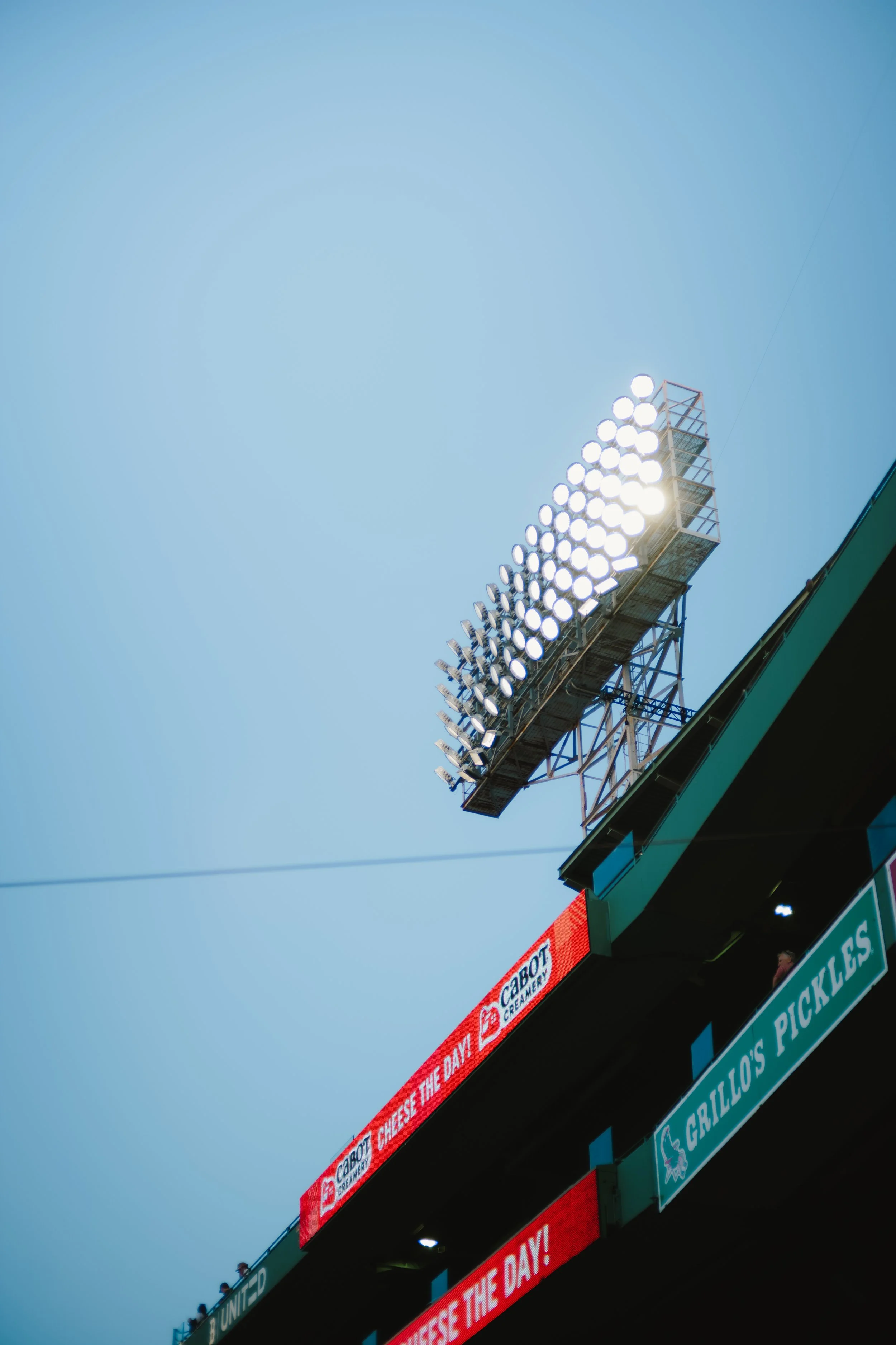 View of a stadium scoreboard with brightly lit lights in the upper right corner and advertising banners in the lower part of the image. The sky is clear and blue.